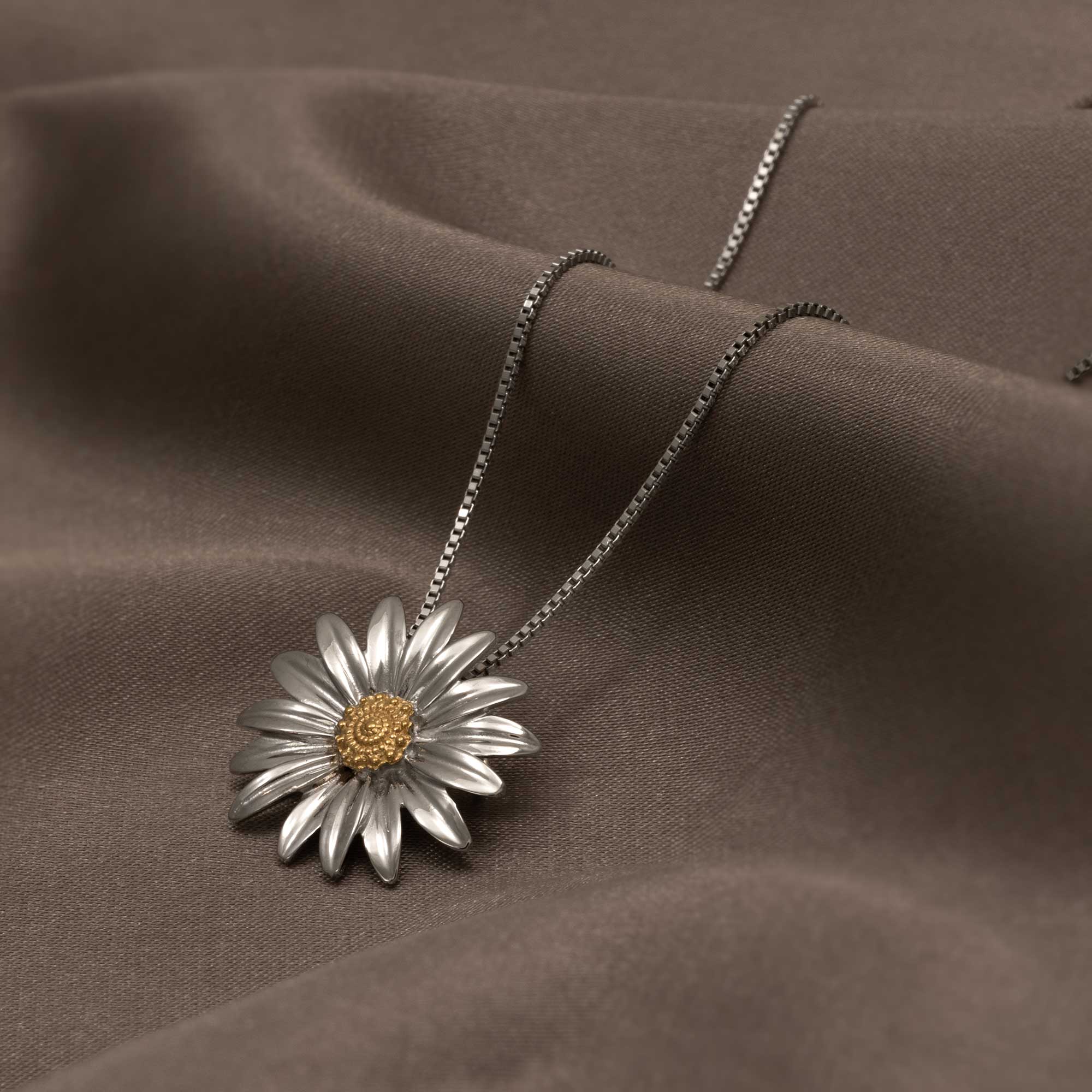 Sterling silver pendant crafted as a detailed daisy flower with a textured gold vermeil center, suspended from a fine chain over smooth brown fabric.