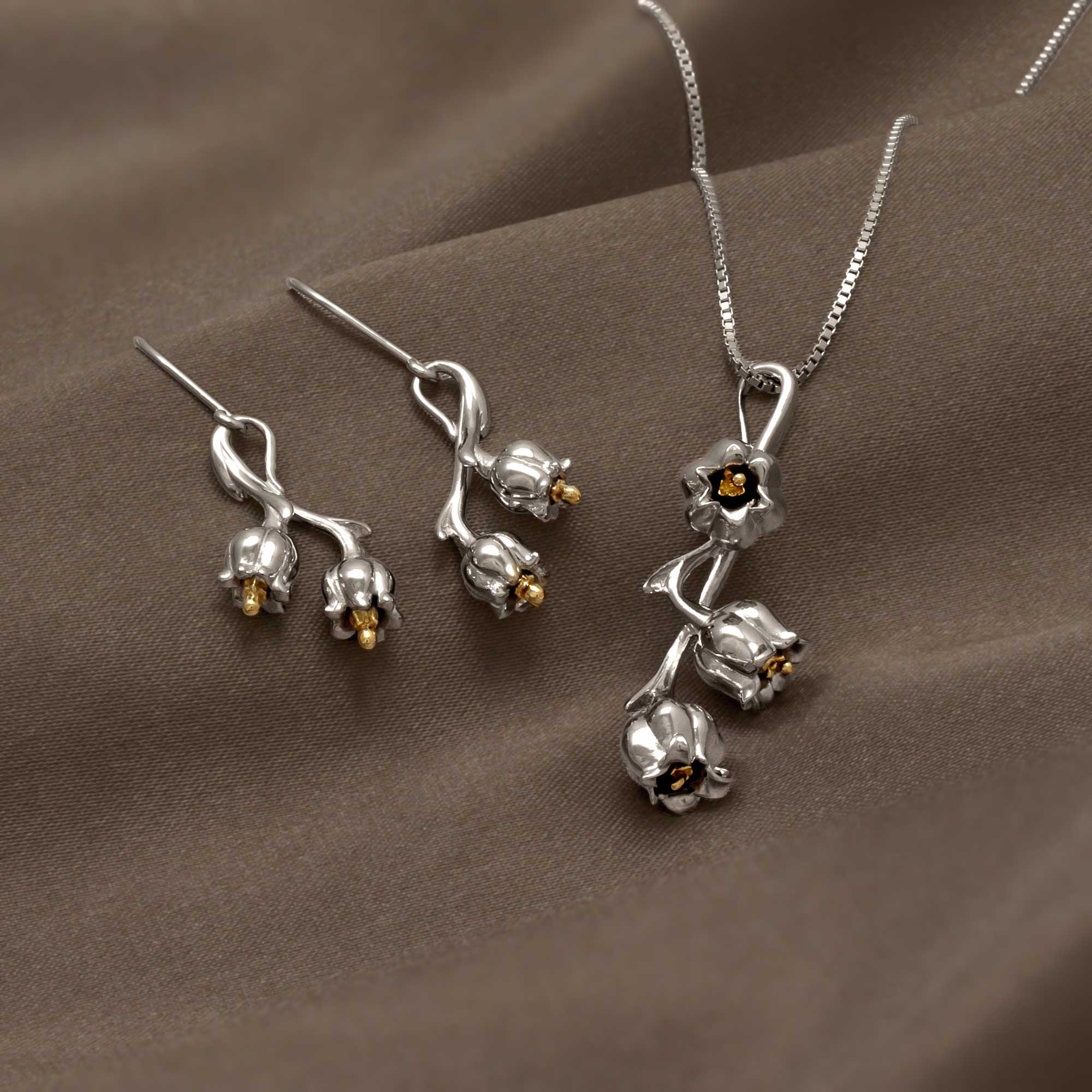 Sterling silver floral necklace and earrings set featuring sculptural Lily of the Valley motifs with delicate gold-tone accents, displayed on soft brown fabric.