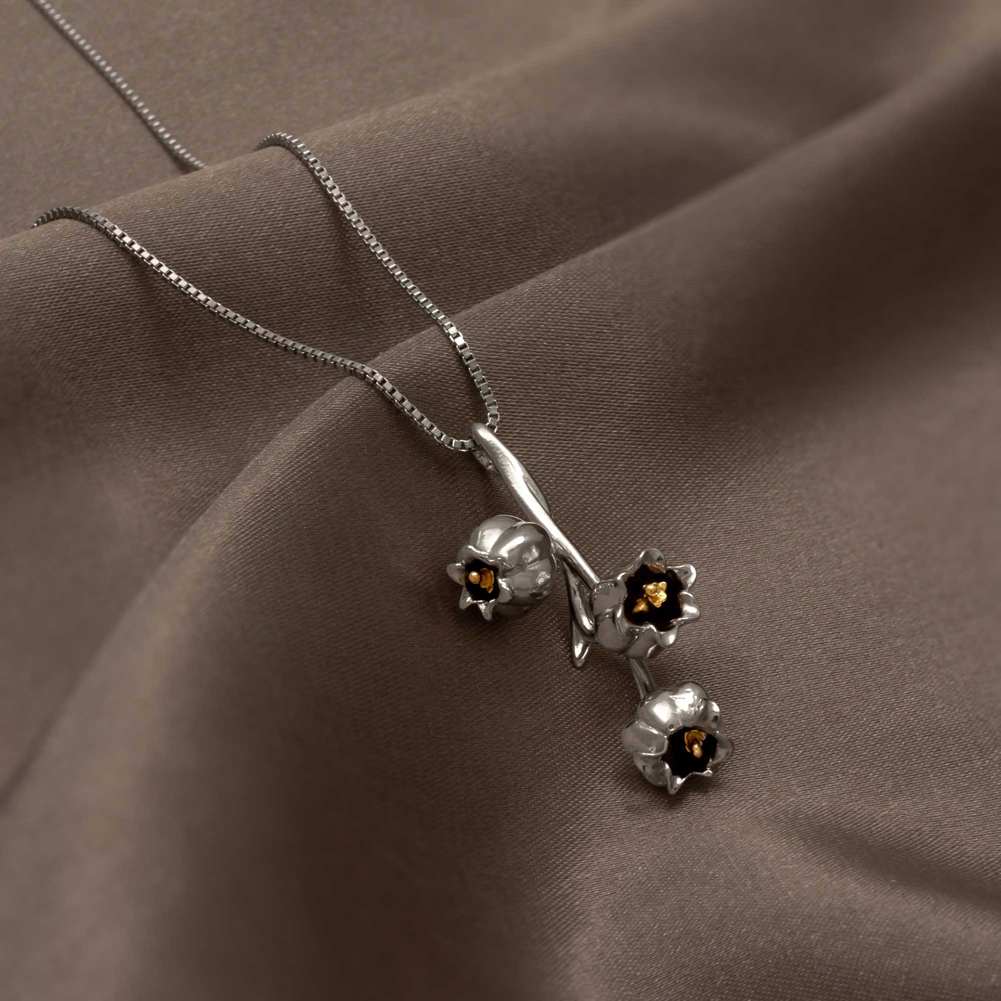 Sterling silver pendant inspired by delicate lily of the valley blossoms with subtle gold accents, suspended on a fine chain against a smooth taupe fabric background.