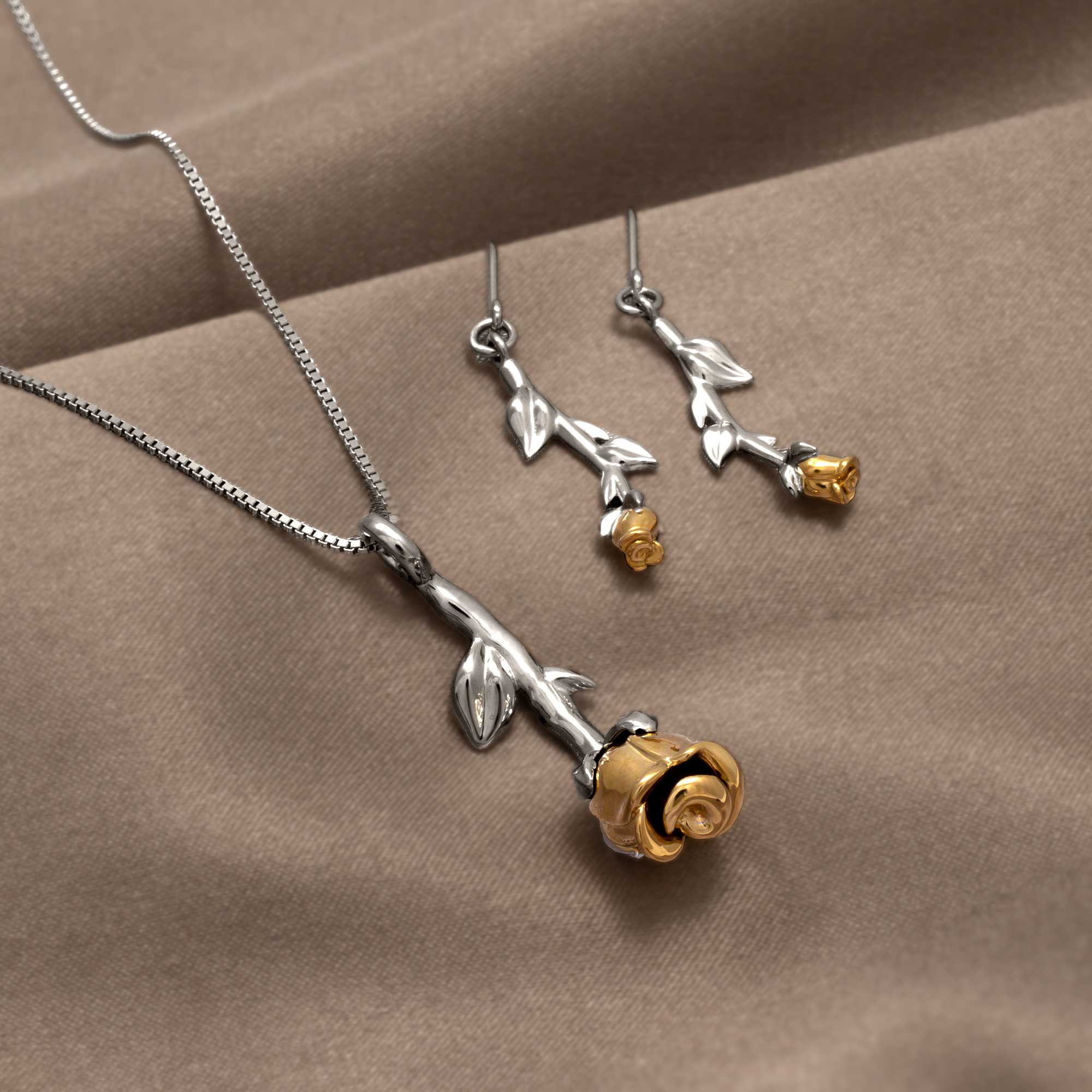 Sterling silver necklace and matching earrings featuring sculptural rose motifs with gold vermeil accents, displayed on a luxe taupe fabric background.