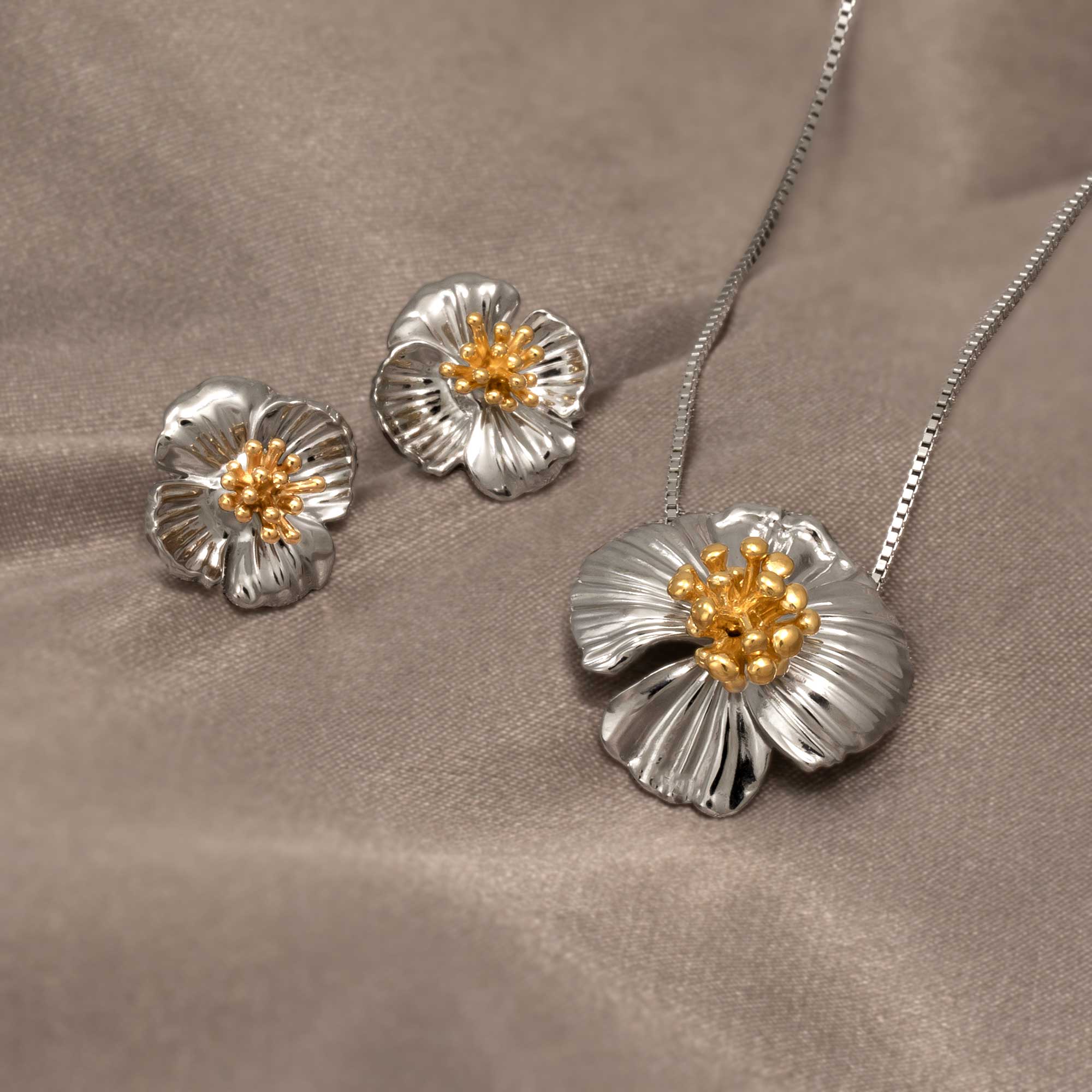 Sterling silver earrings and matching pendant featuring sculpted poppy flower designs with textured petals and gold-tone stamen details, displayed on a soft taupe fabric.