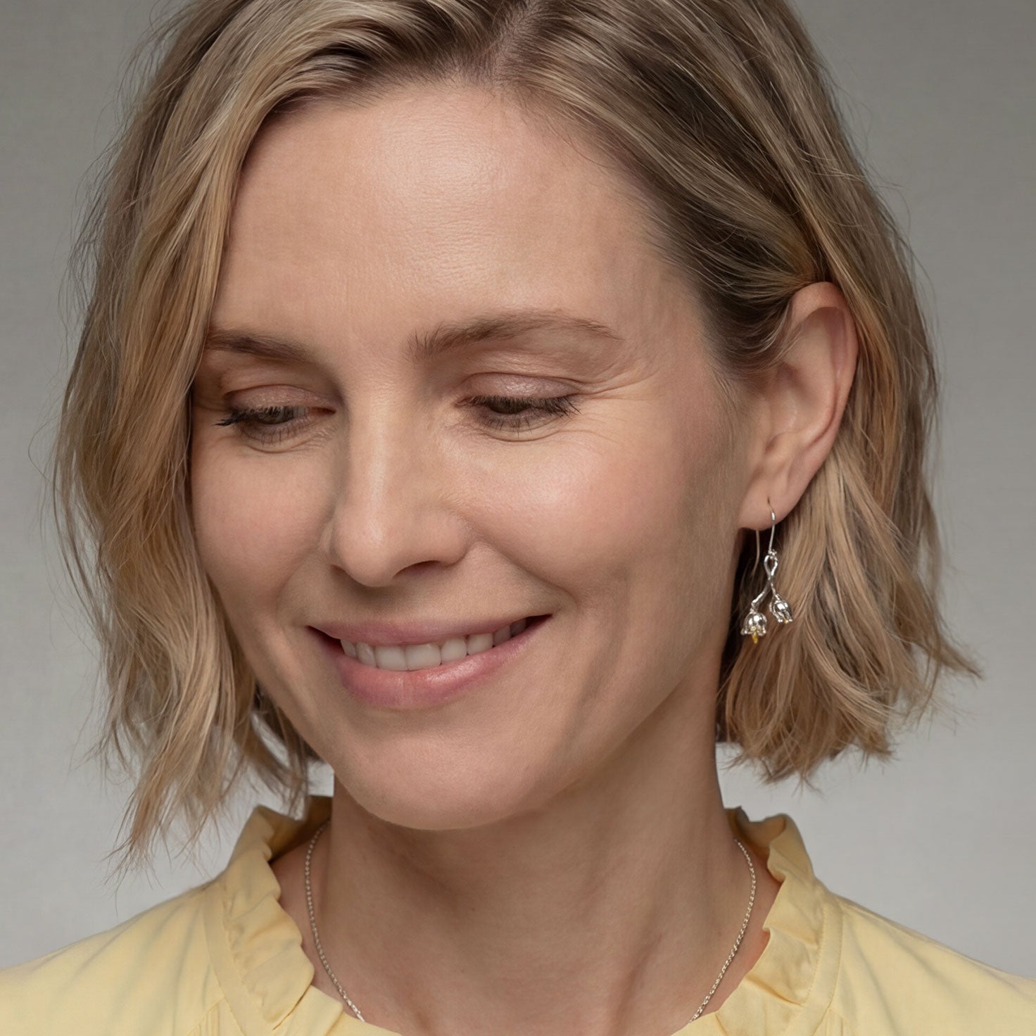 Delicate sterling silver earrings inspired by lily of the valley flowers, featuring sculptural floral charms suspended from slender hook clasps, worn with a soft smile.