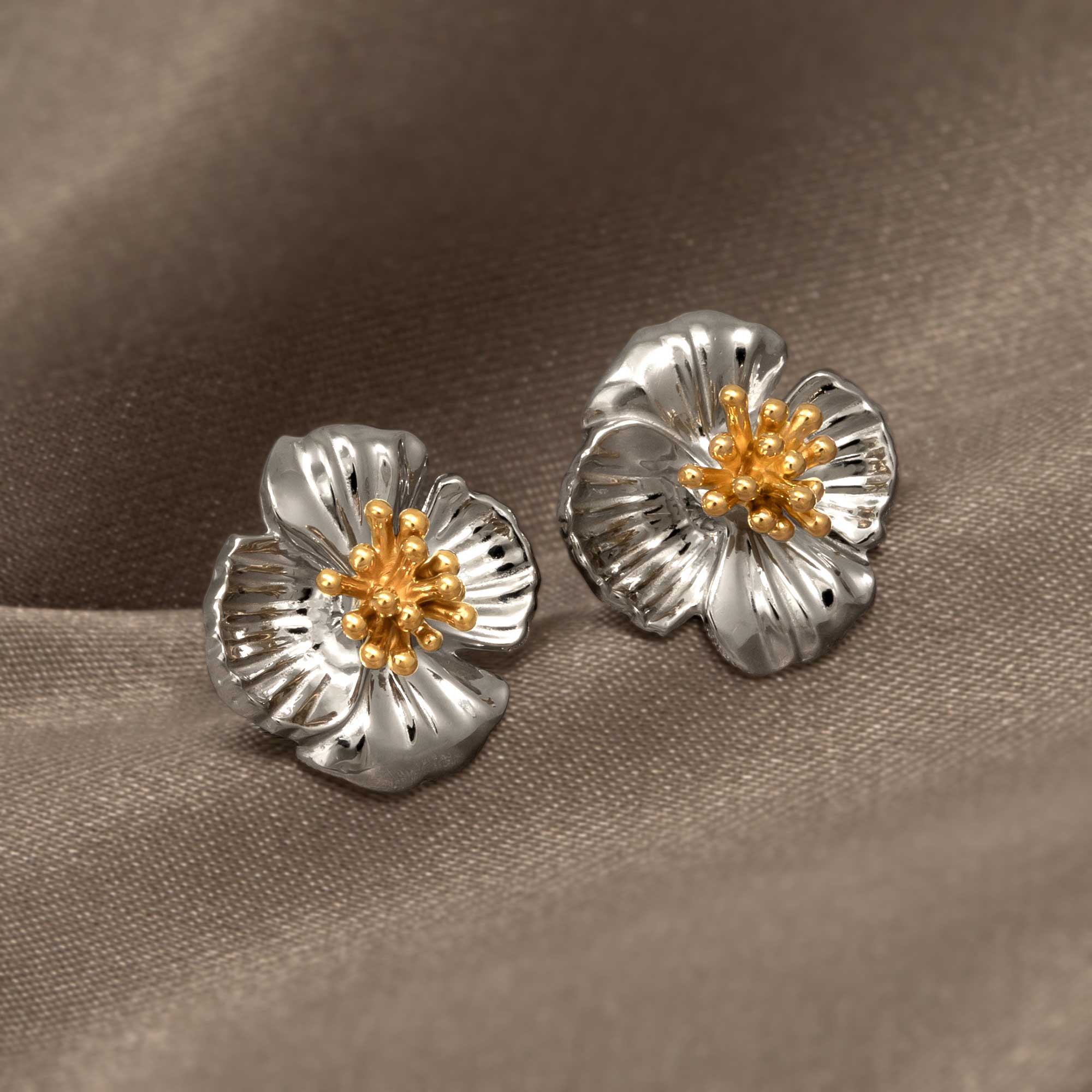 Sterling silver flower-shaped earrings with sculptural petals accented by gold vermeil detailing, presented on a smooth taupe fabric background.