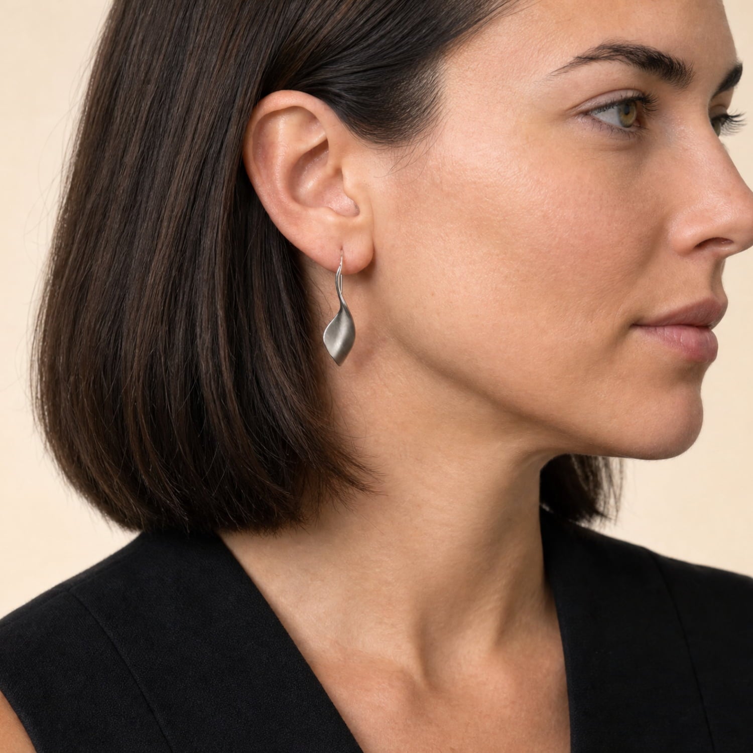 A close-up side profile of a woman with dark brown hair wearing a twisting drop earring, set against a plain beige background.
