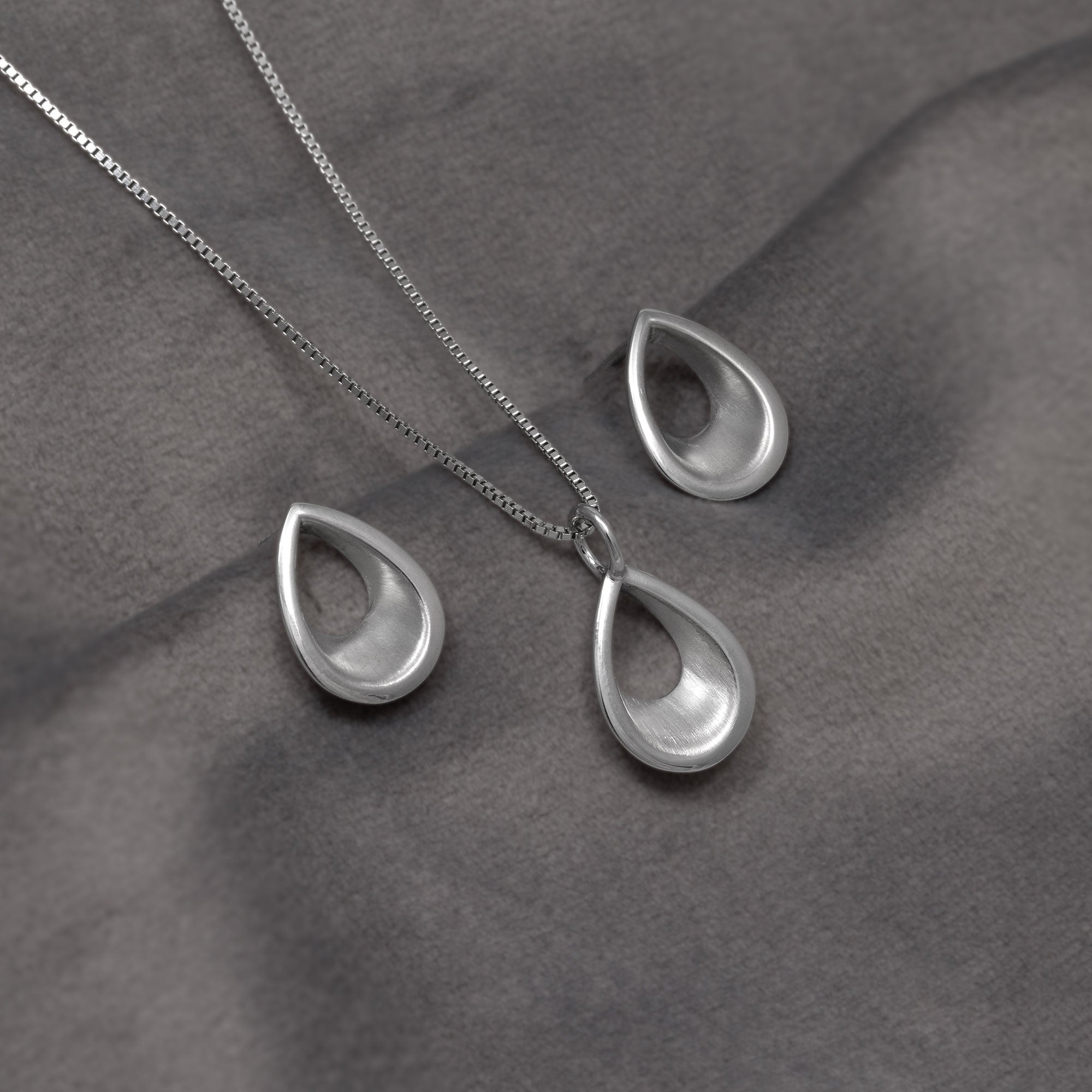 Silver teardrop pendant necklace and matching drop earrings on a dark grey fabric background.