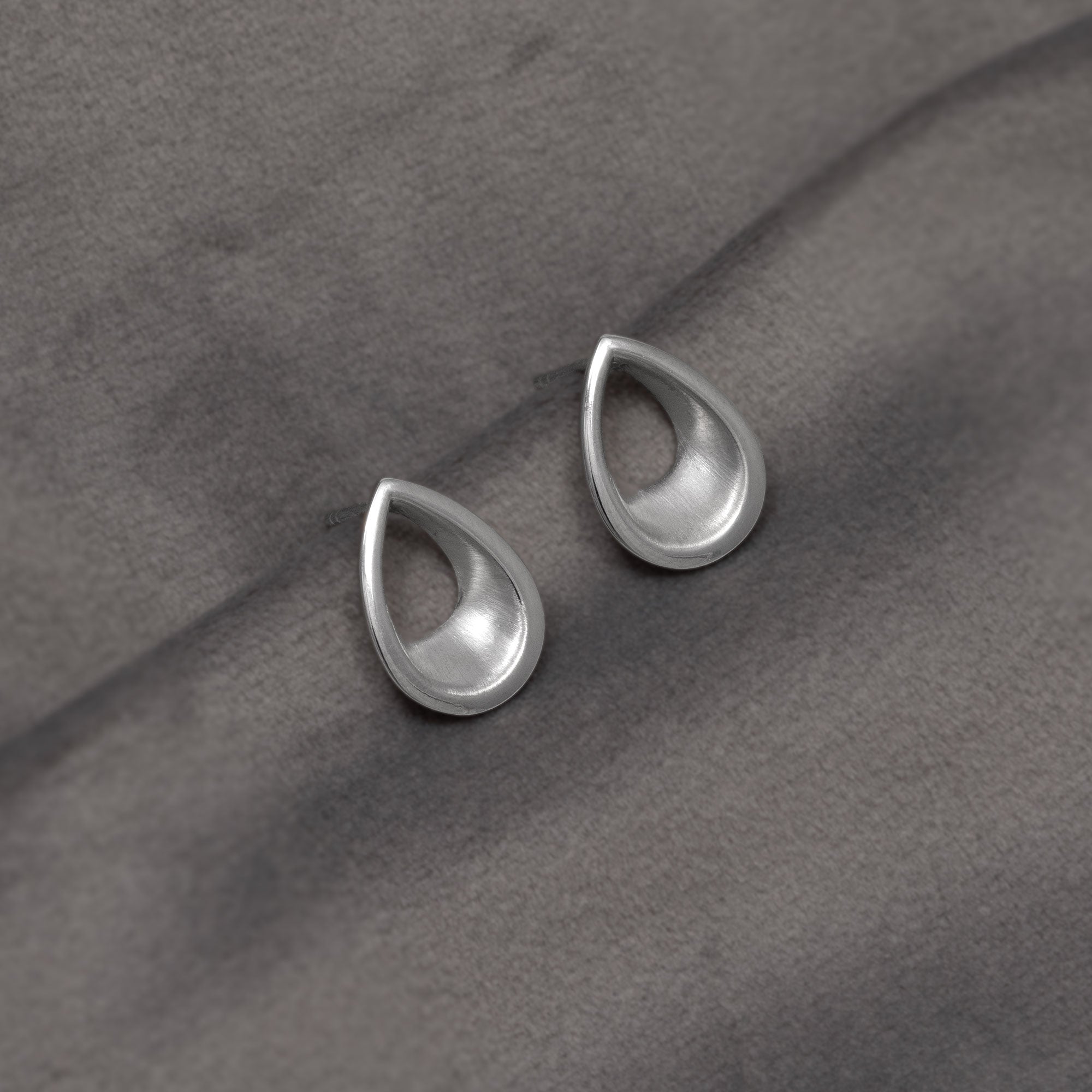 A pair of silver teardrop-shaped stud earrings with a polished, open centre design, resting on a textured dark grey fabric background.