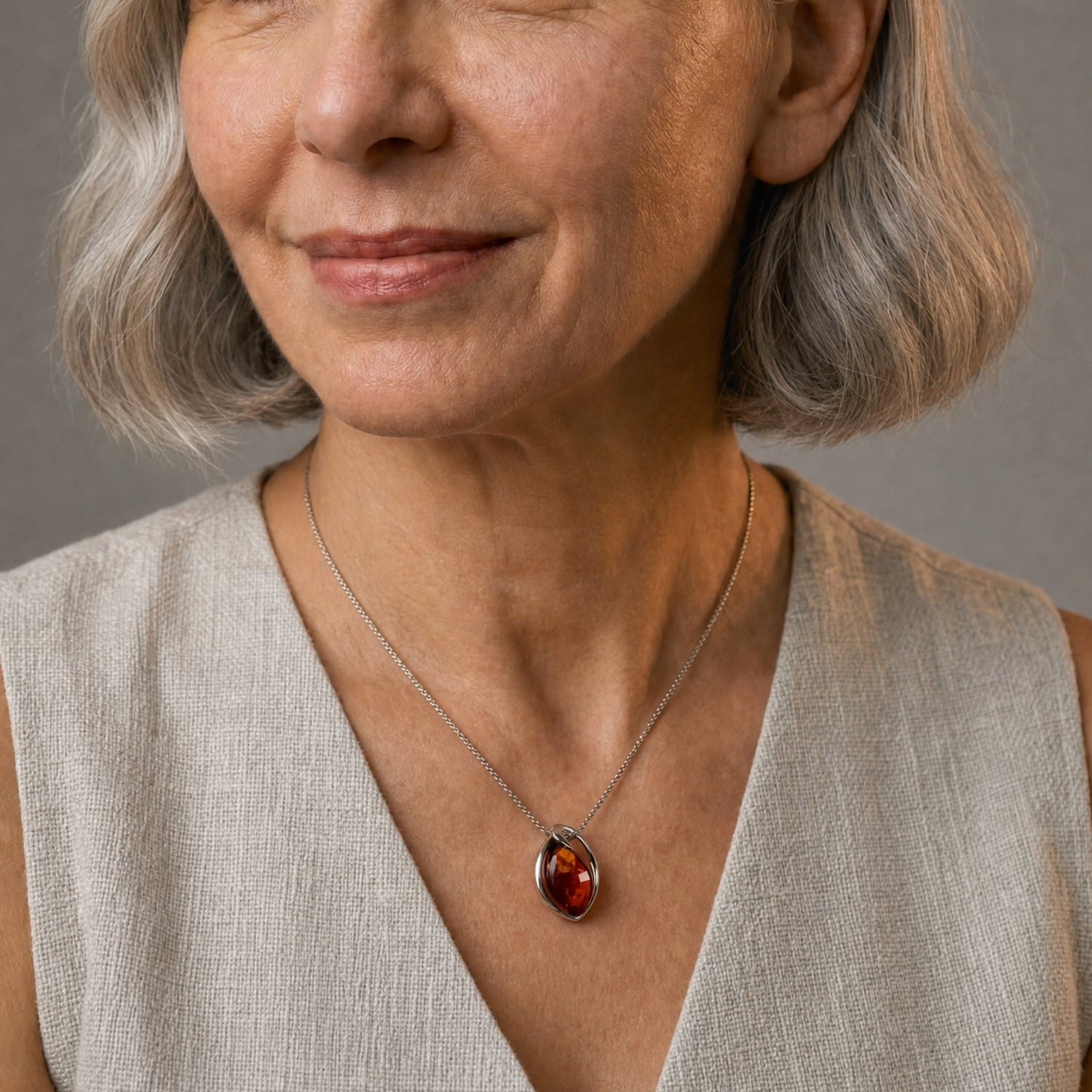 Close-up of a woman wearing a silver chain necklace with an oval amber gem, shown against a soft gray background.