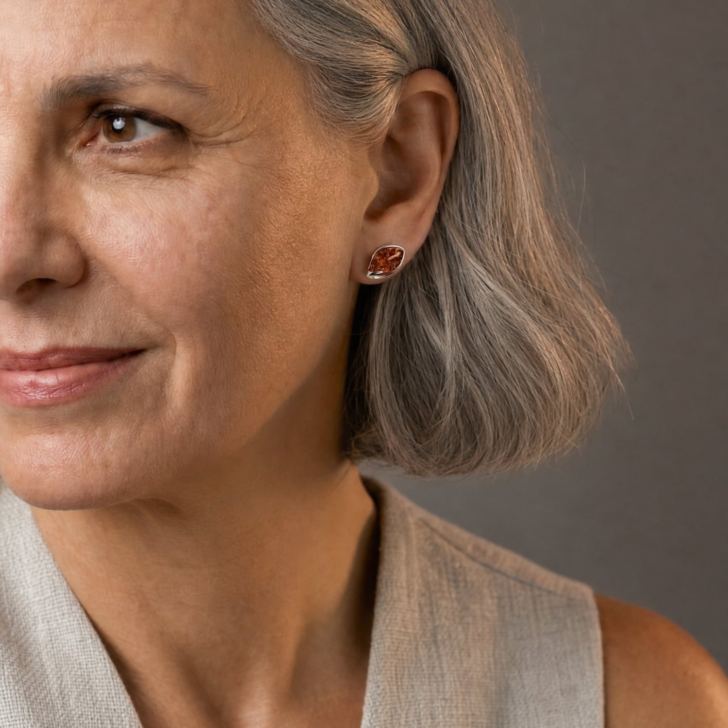 Close-up of a woman with grey hair wearing a small, organic-shaped amber stud earring, shown against a neutral gray background.
