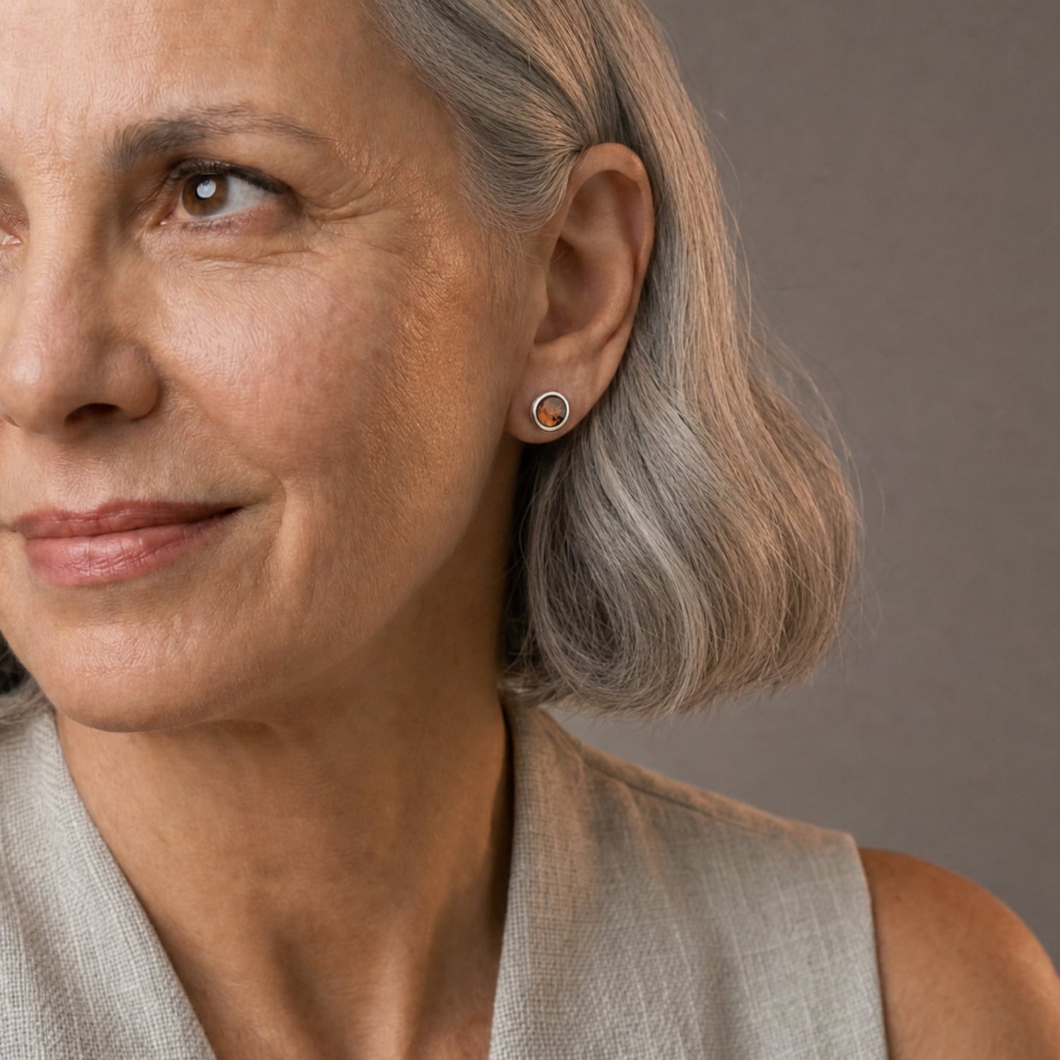 Close-up of a woman with grey hair wearing a small round stud earring with a central amber gem, against a plain taupe background.