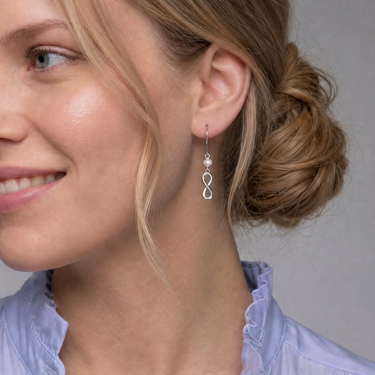 A close-up of a woman wearing a silver infinity drop earring with a small pearl accent, shown against a neutral gray background.