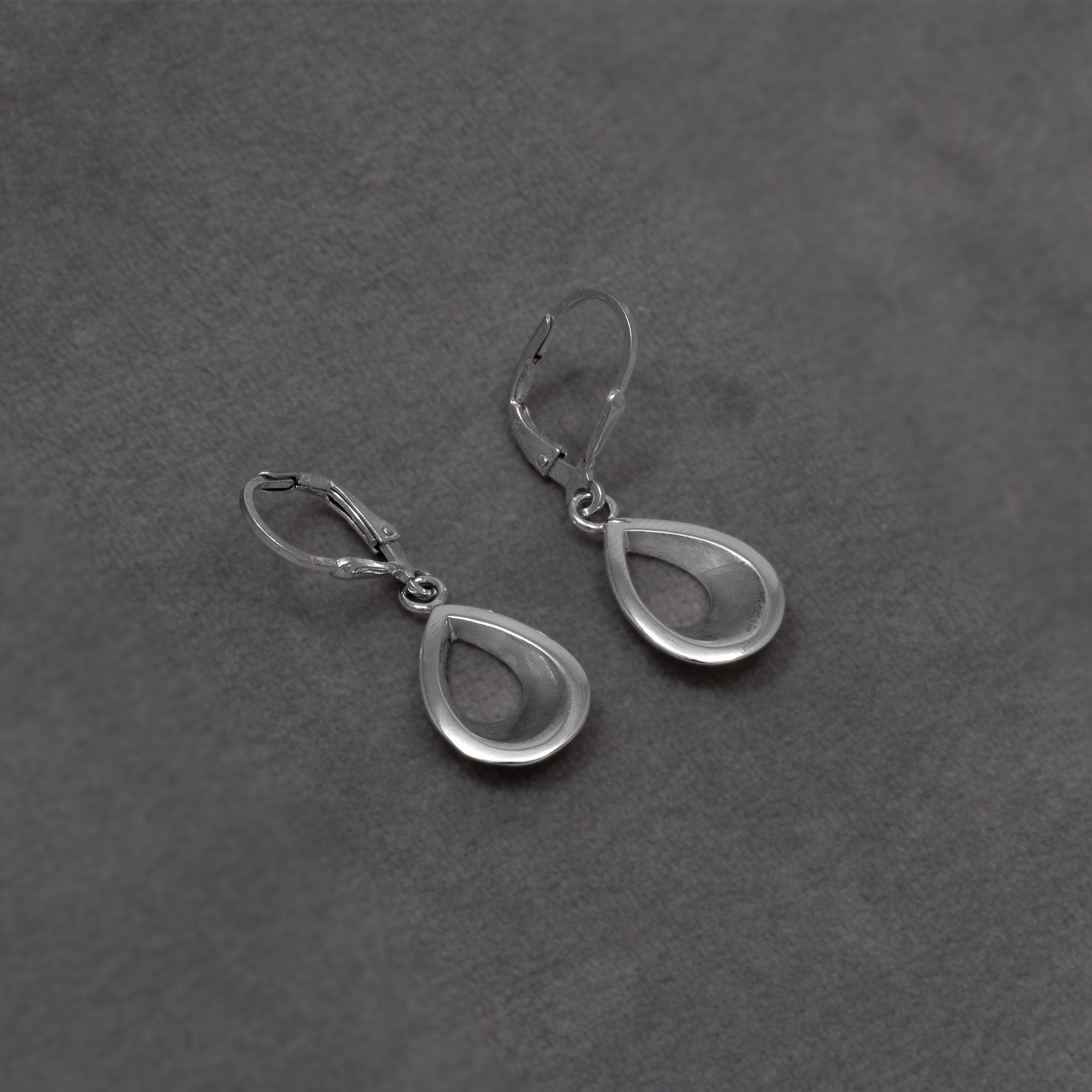 A pair of silver teardrop-shaped drop earrings with leverback closures, displayed on a textured dark grey background.
