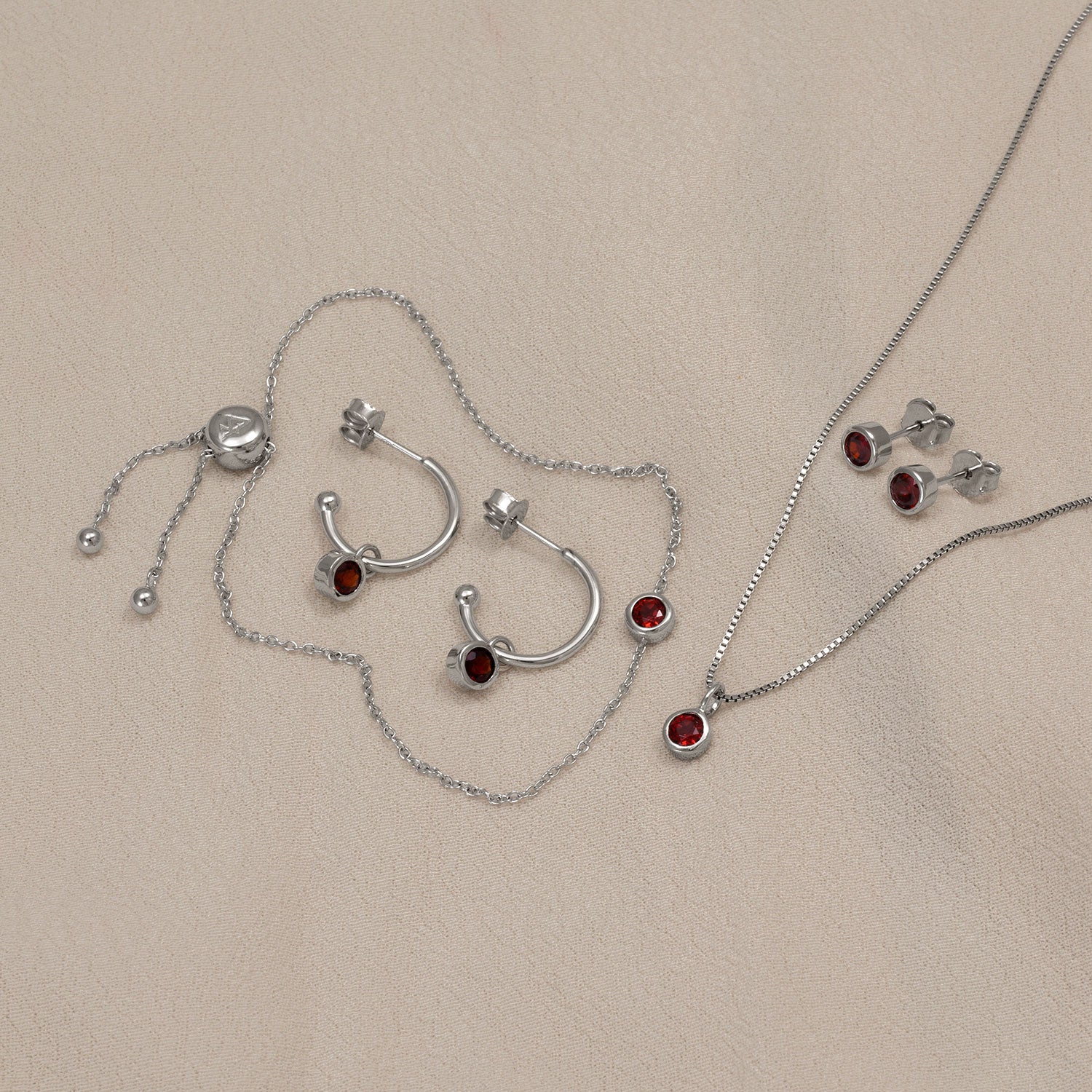 Sterling silver hoop earrings and stud earrings set with deep red garnet stones, displayed alongside a delicate garnet pendant necklace on a soft beige fabric.