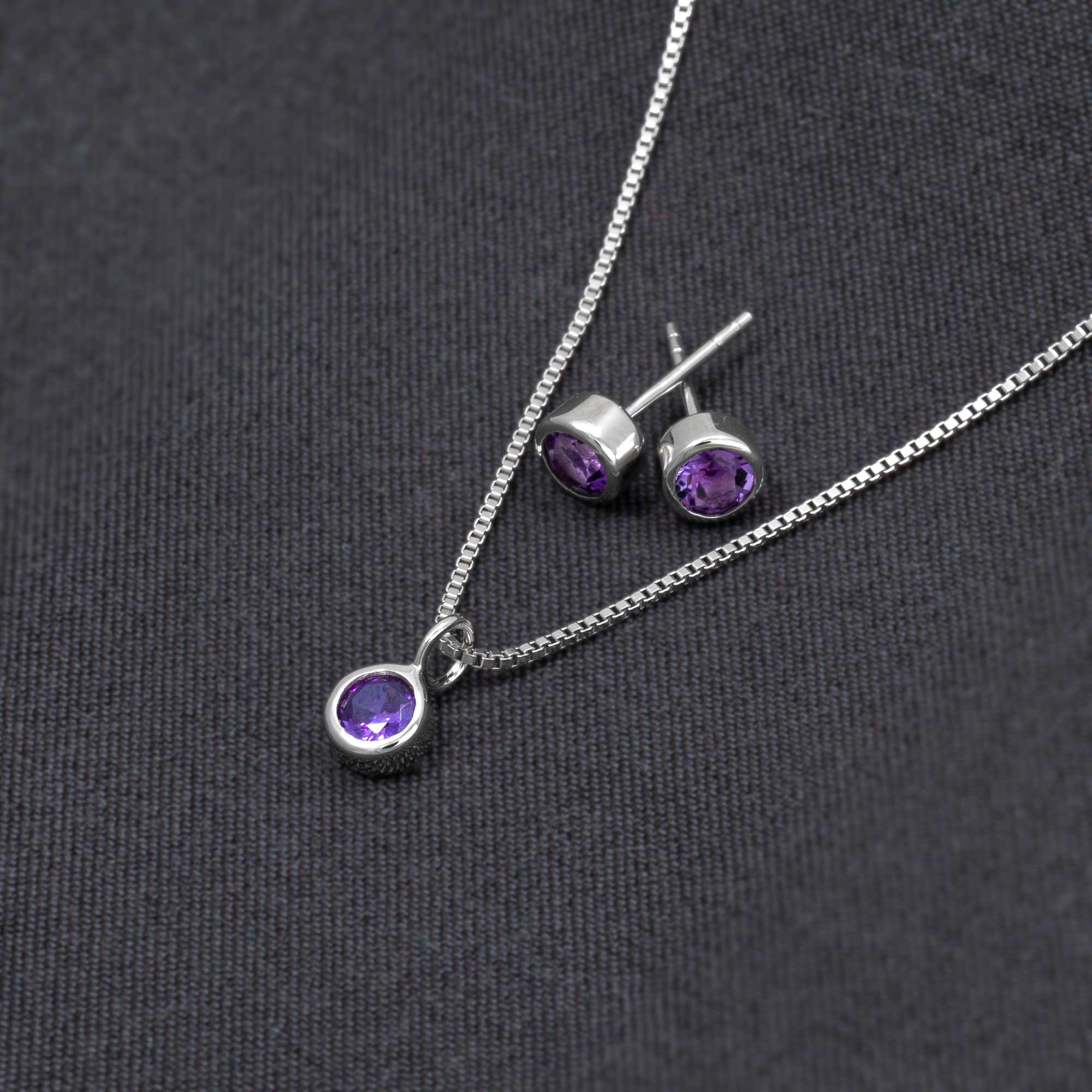 Pair of sterling silver stud earrings with bezel-set amethyst gemstones, accompanied by a matching pendant on a delicate silver chain, displayed on dark fabric.