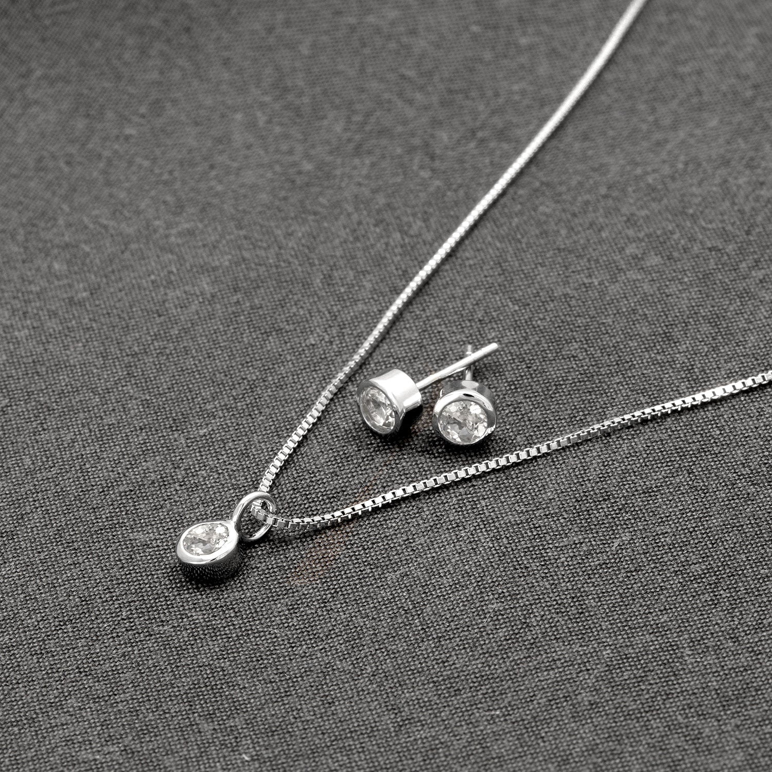 Delicate sterling silver stud earrings and matching pendant featuring bezel-set white topaz gemstones, arranged on a textured dark grey fabric background.