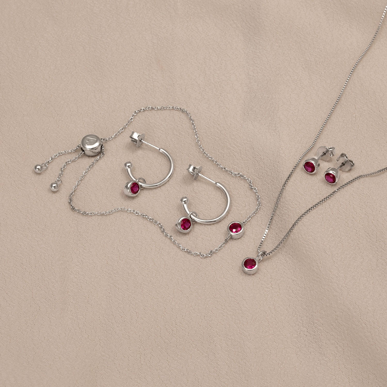 Sterling silver jewellery set featuring hoop earrings, a pendant necklace, and stud earrings each adorned with vibrant ruby gemstones, presented on a neutral fabric background.
