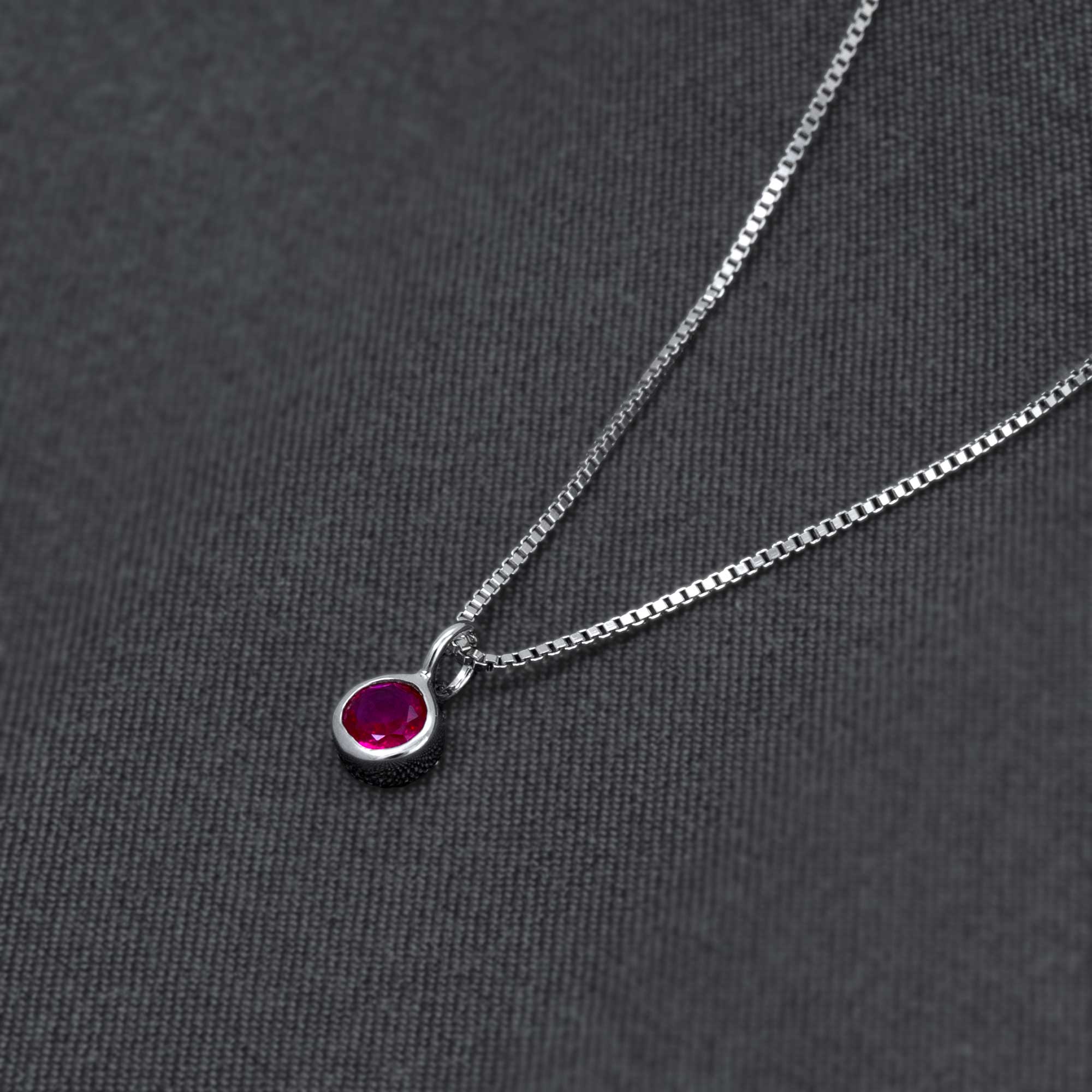 Delicate sterling silver pendant featuring a vibrant ruby birthstone set in a minimalist bezel design, suspended from a fine chain on a dark textured background.