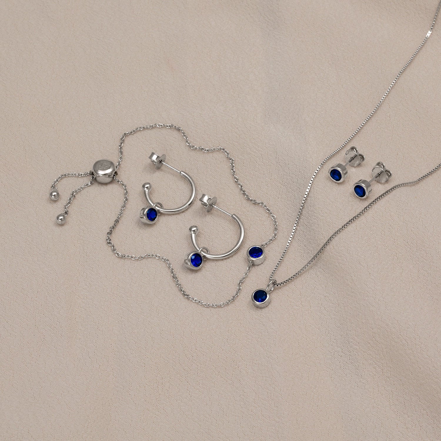 Sterling silver jewellery set featuring deep blue spinel stones, including hoop earrings, stud earrings, and a pendant necklace, displayed on a neutral fabric.