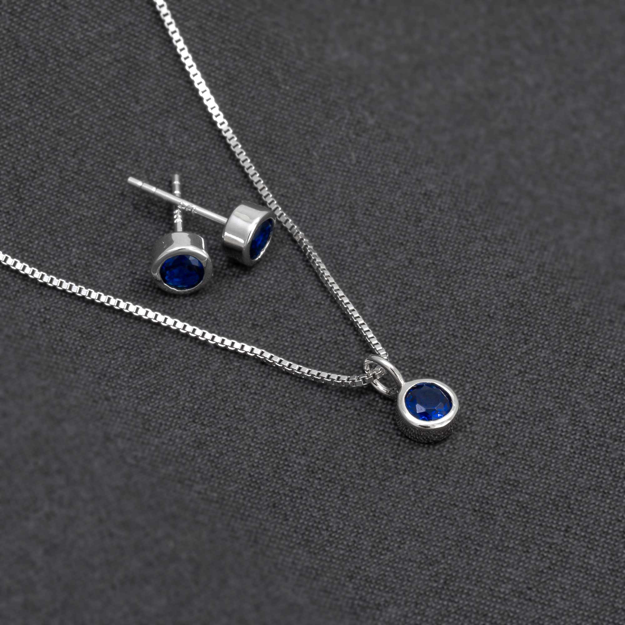 Sterling silver stud earrings and a matching pendant set featuring deep blue spinel gemstones, displayed on a dark textured fabric background emphasizing refined craftsmanship.