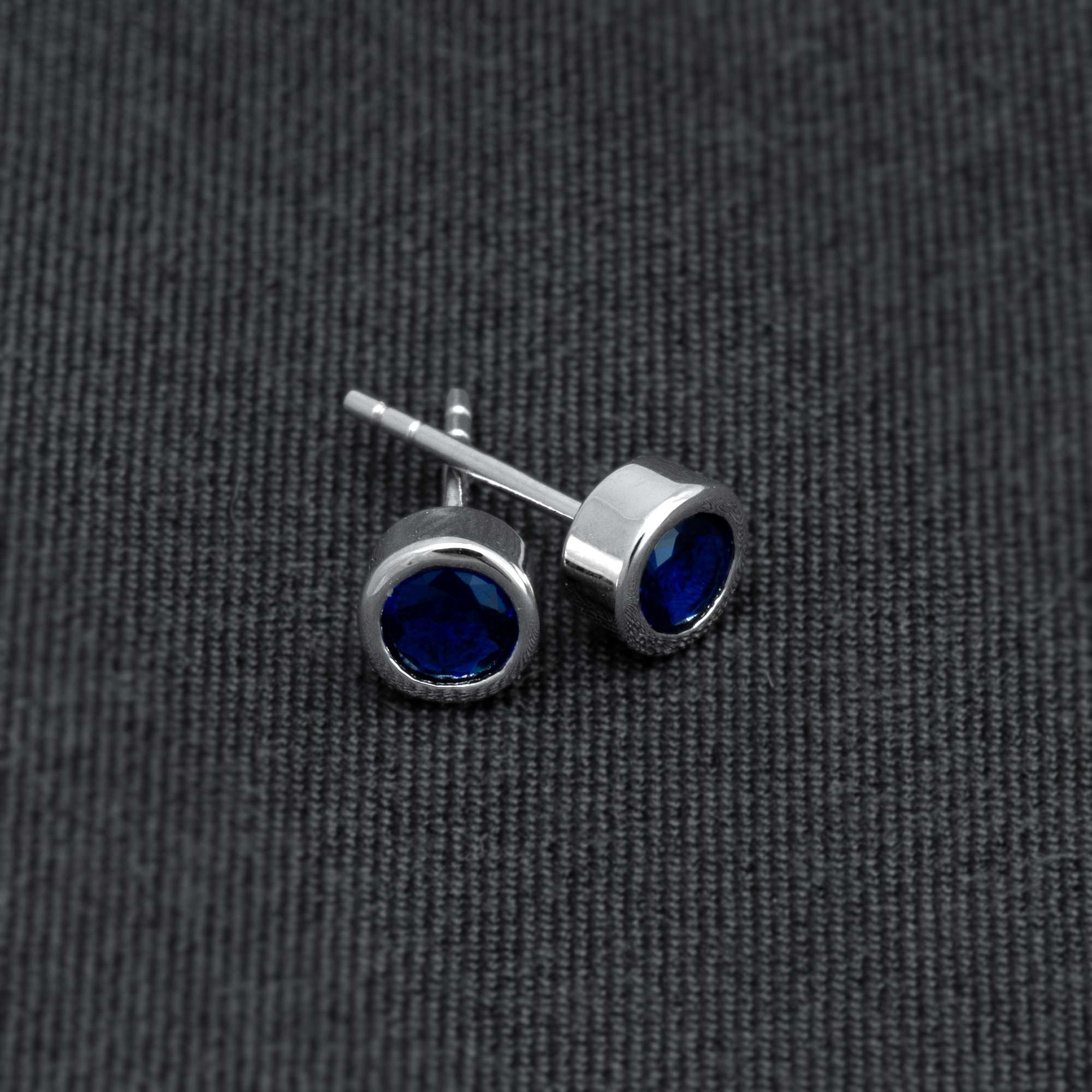 Pair of sleek sterling silver stud earrings set with deep blue spinel gemstones, showcasing refined craftsmanship and a modern, minimalist design on a dark fabric backdrop.