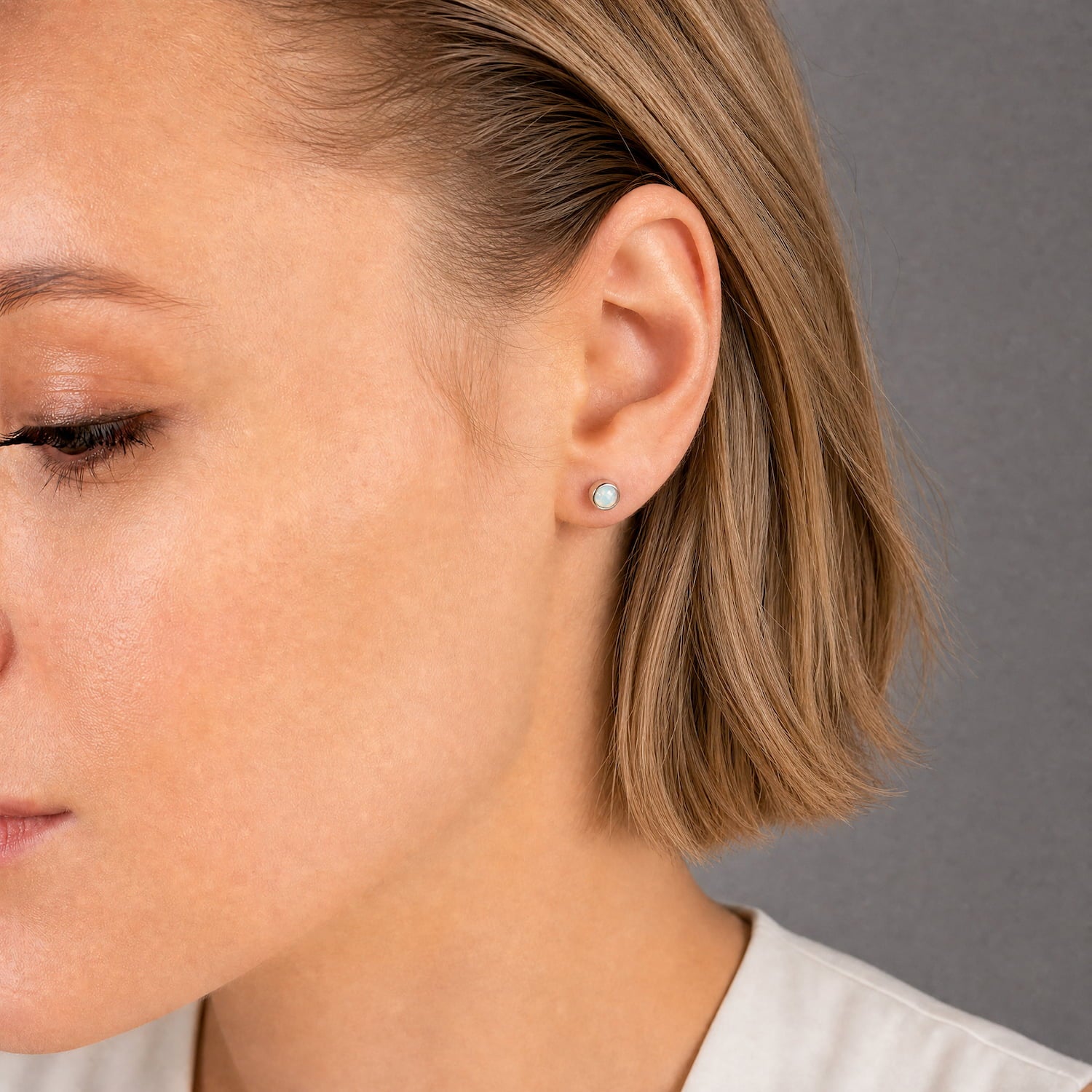 Model wears a pair of sterling silver stud earrings featuring round opal gemstones with a polished bezel setting, highlighting the iridescent play of colors against a dark fabric background.