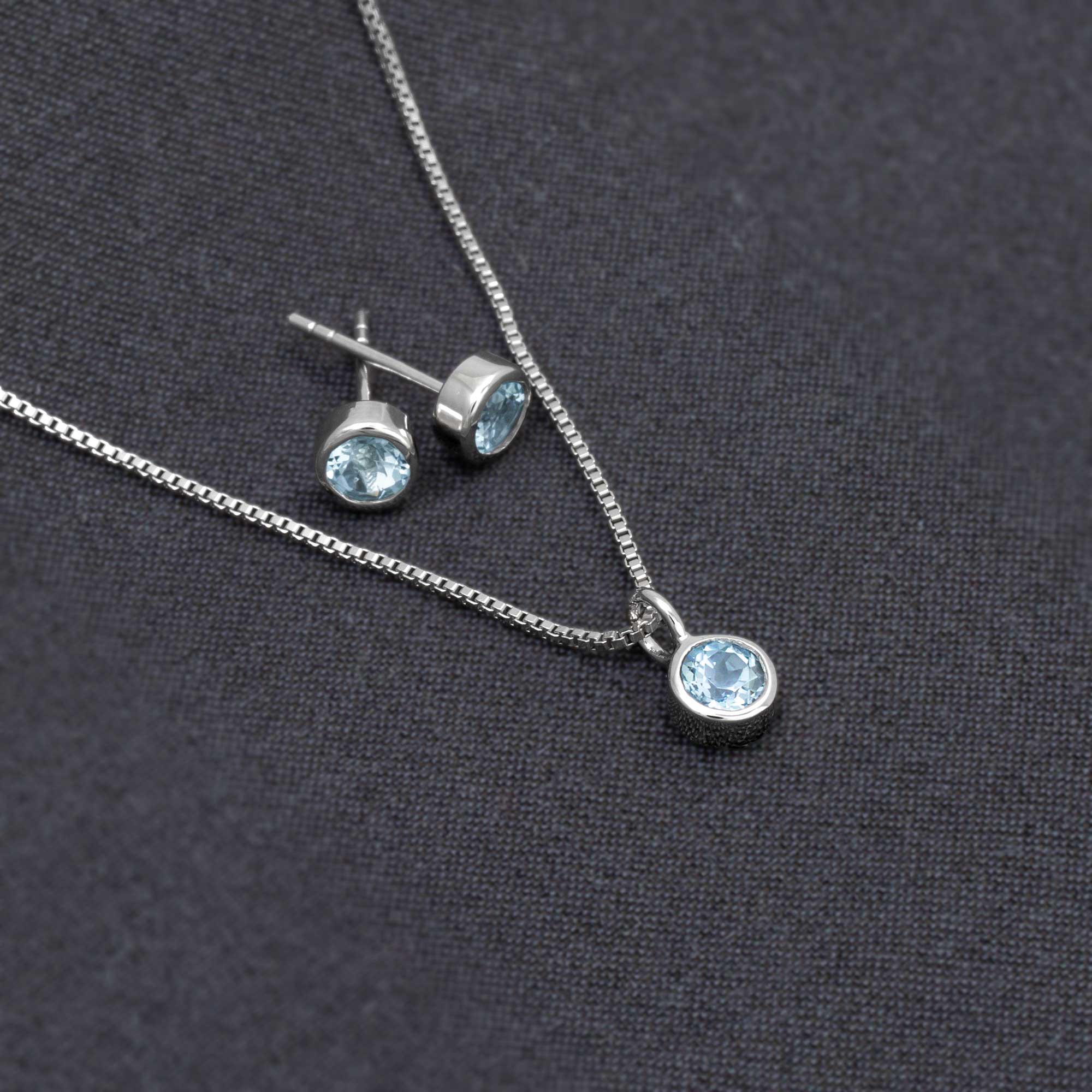 Sterling silver earrings and pendant set featuring vibrant blue topaz gemstones, presented on a fine chain and set against a dark textured fabric background.