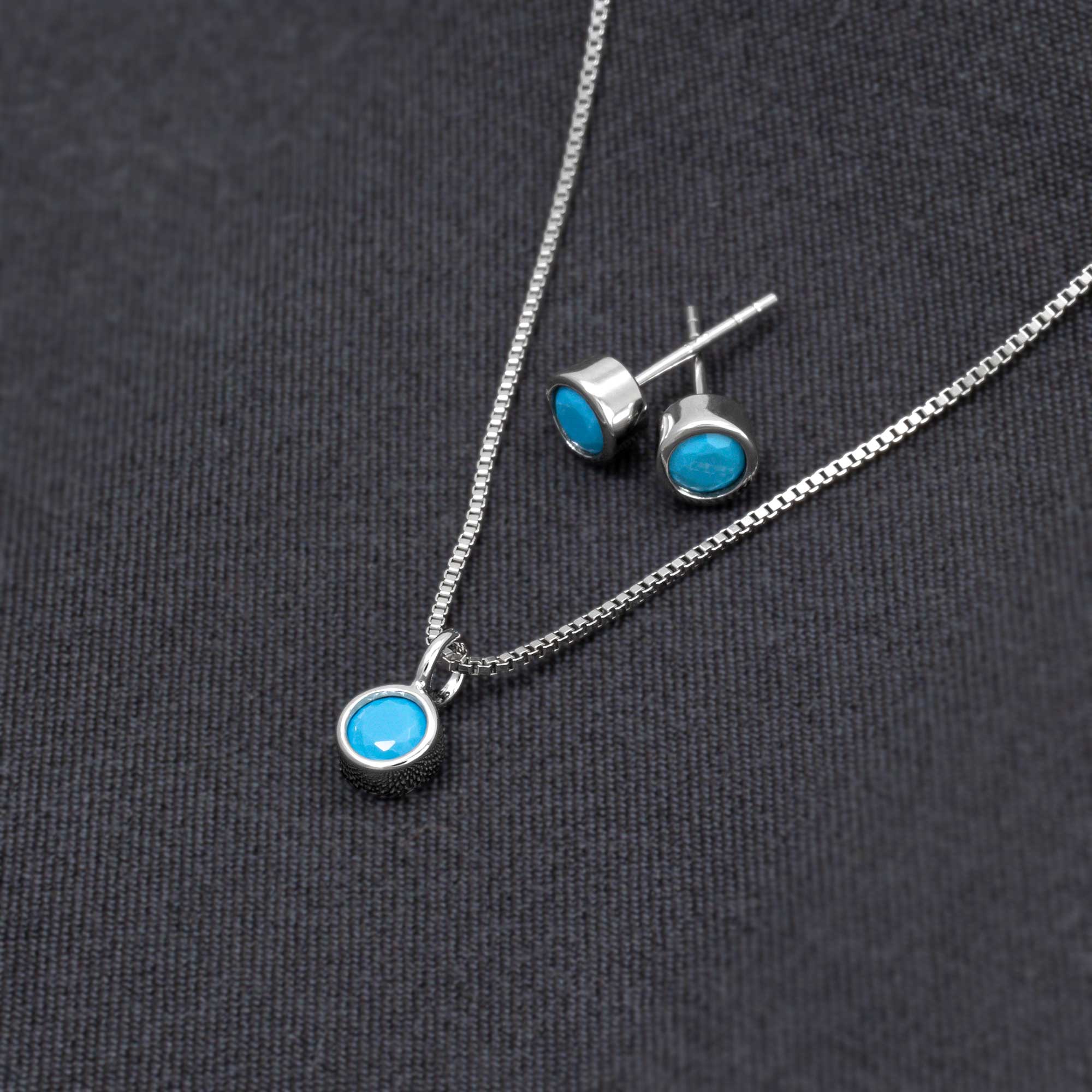 Sterling silver stud earrings showcasing bright turquoise gemstones, designed as contemporary birthstone jewellery with a polished finish, set against a dark fabric background.