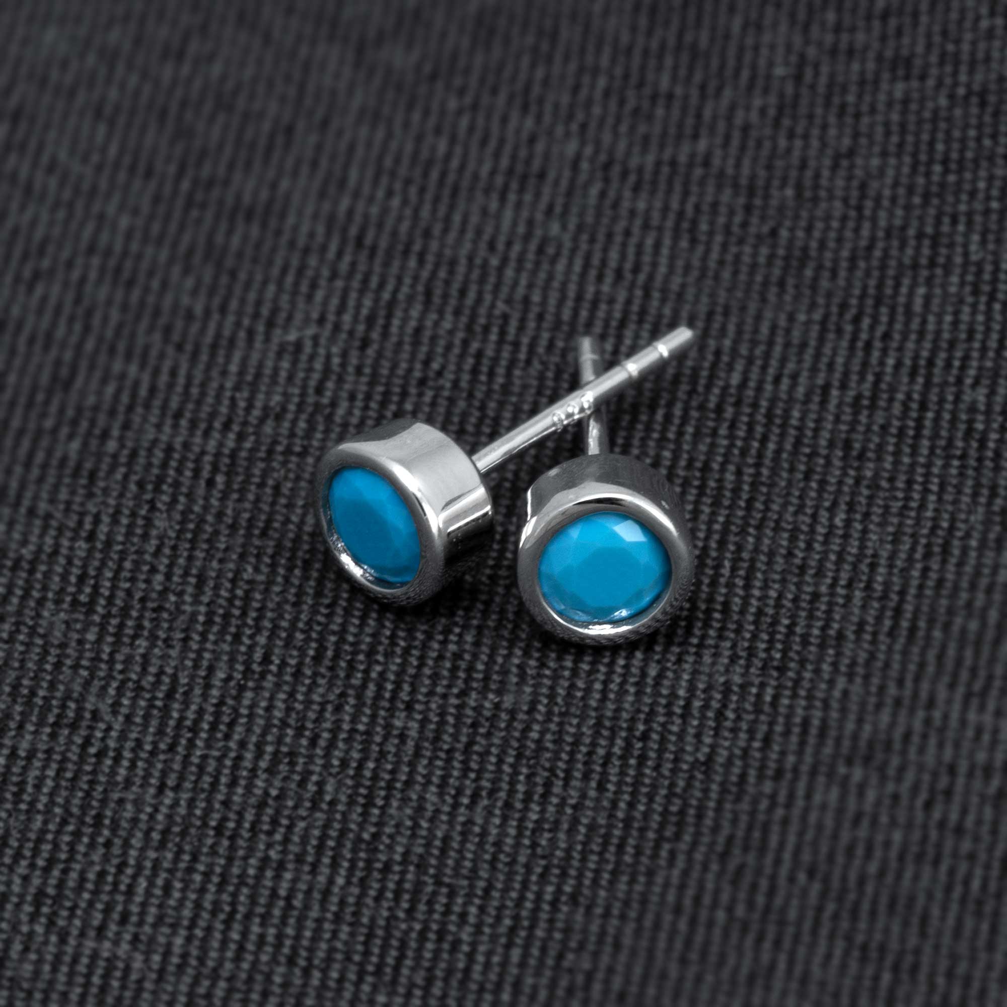Pair of sterling silver stud earrings featuring round turquoise gemstones set in a polished bezel, presented on a dark textured fabric background.