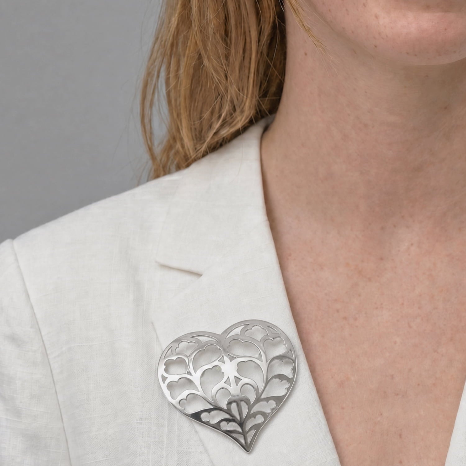 A large sterling silver brooch shaped as an openwork heart with intricate leaf-inspired detailing, styled in a white linen jacket.