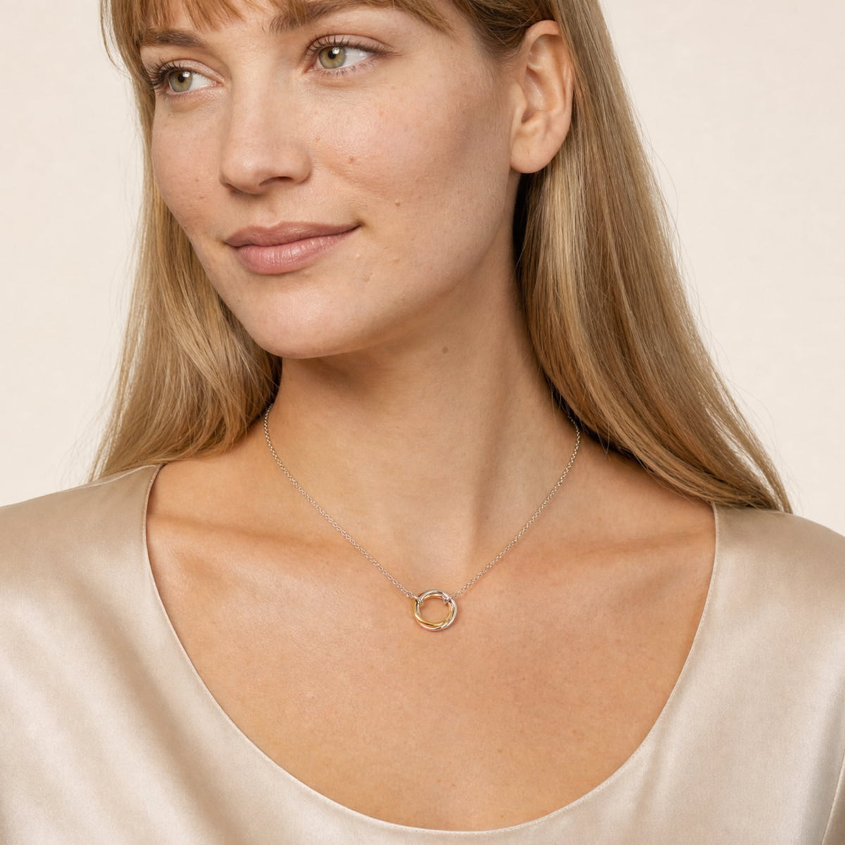 A close-up of a woman wearing a silver chain necklace with a triple-ring pendant. She is wearing a satin champagne-colored top against a soft, light beige background.