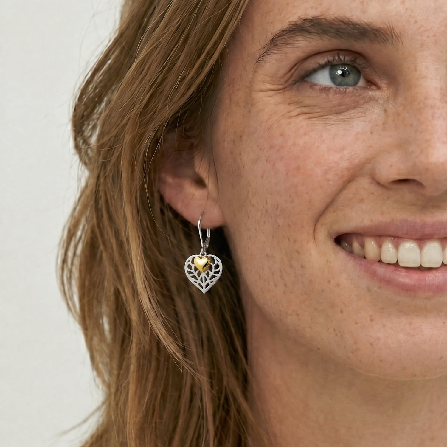 Close-up of a model wearing a double drop earring with a gold vermeil heart charm suspended inside a delicate sterling silver openwork heart, showcasing expert craftsmanship.