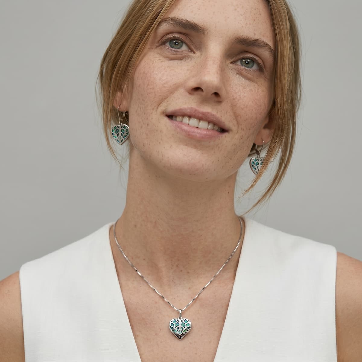 Woman wearing sterling silver earrings and matching pendant with intricate heart-shaped design featuring abalone inlay, styled with a simple white top.