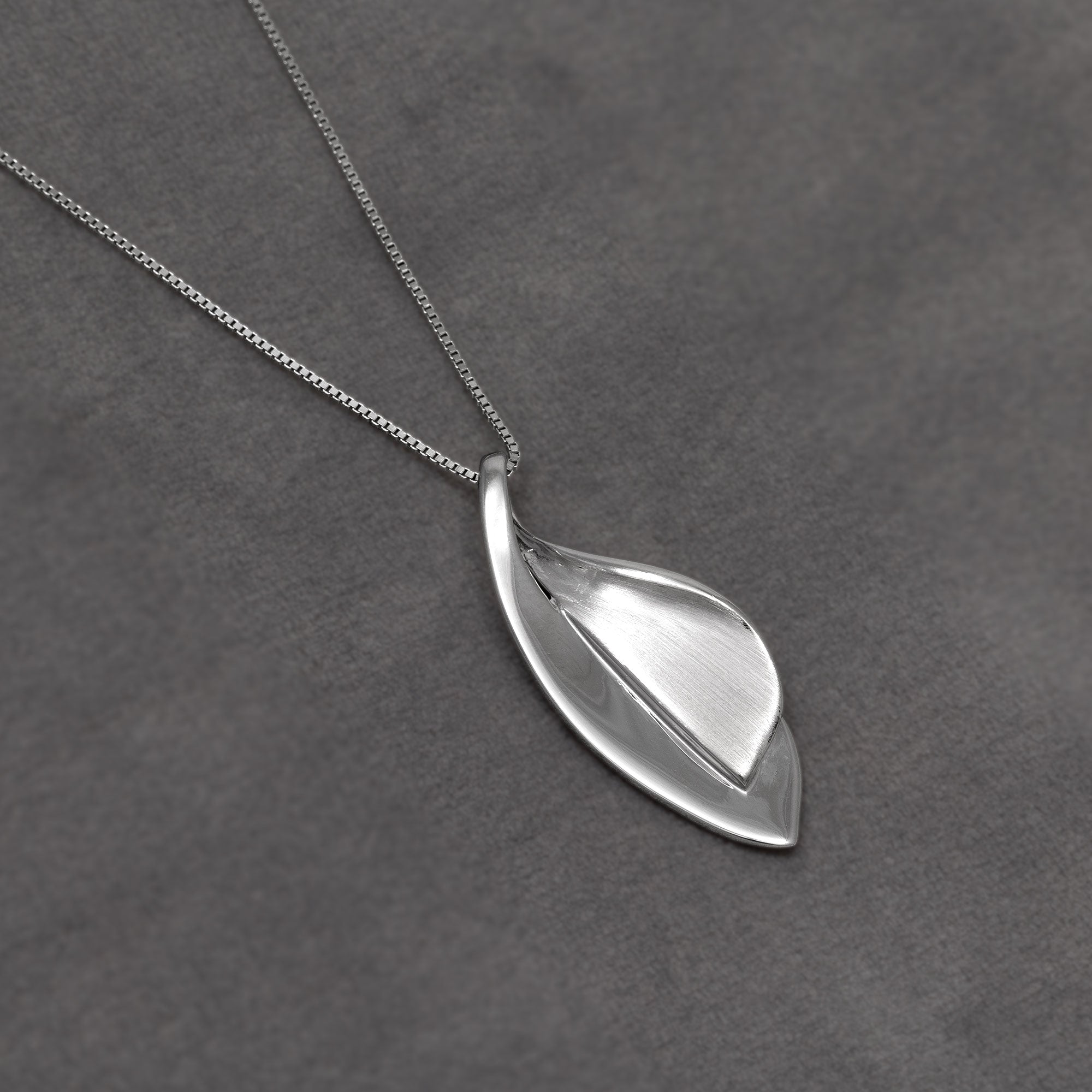 A silver pendant necklace featuring a stylized leaf design with a brushed centre and polished side, resting on a textured dark grey surface.