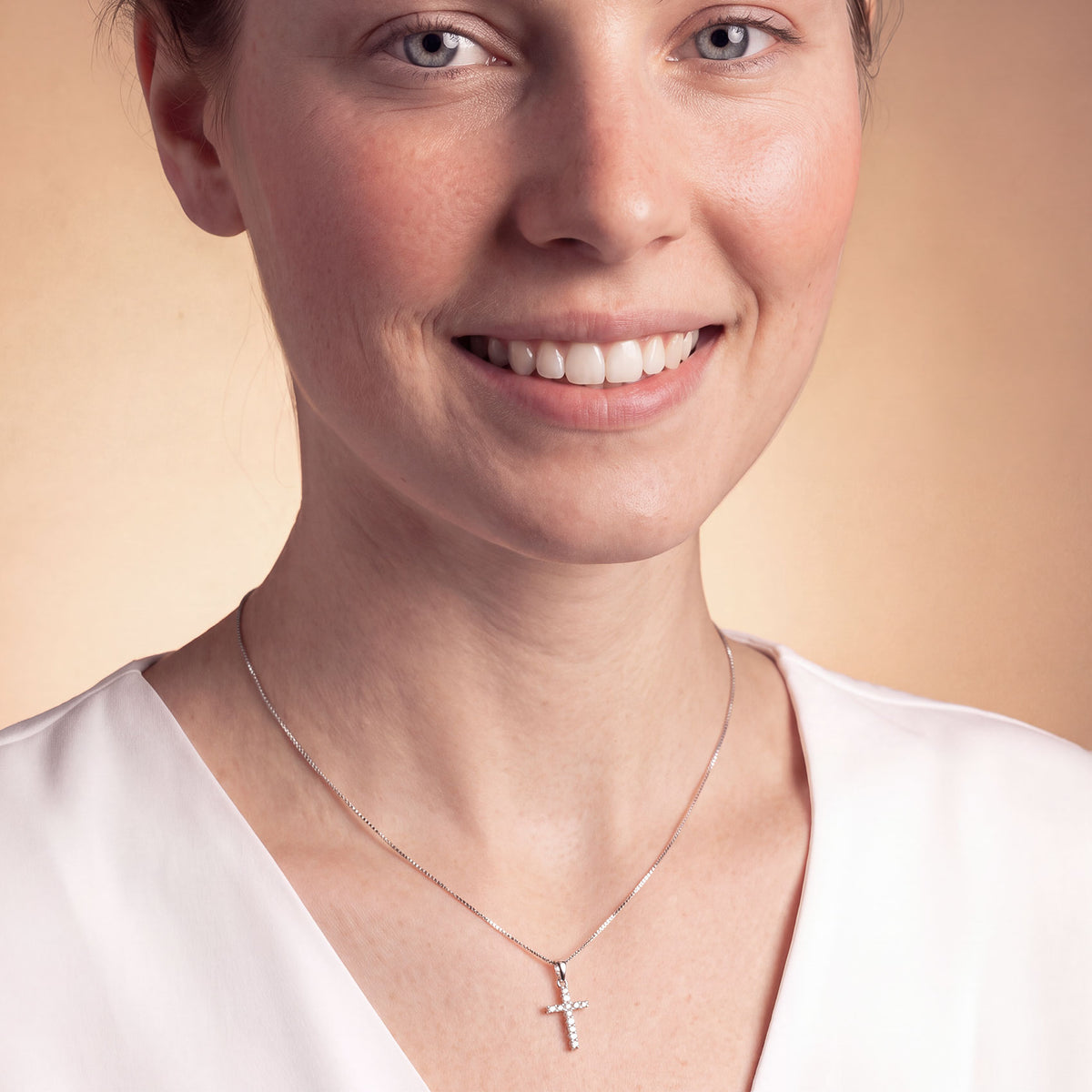 A close-up of a smiling woman wearing a delicate silver cross pendant necklace on a warm beige background.