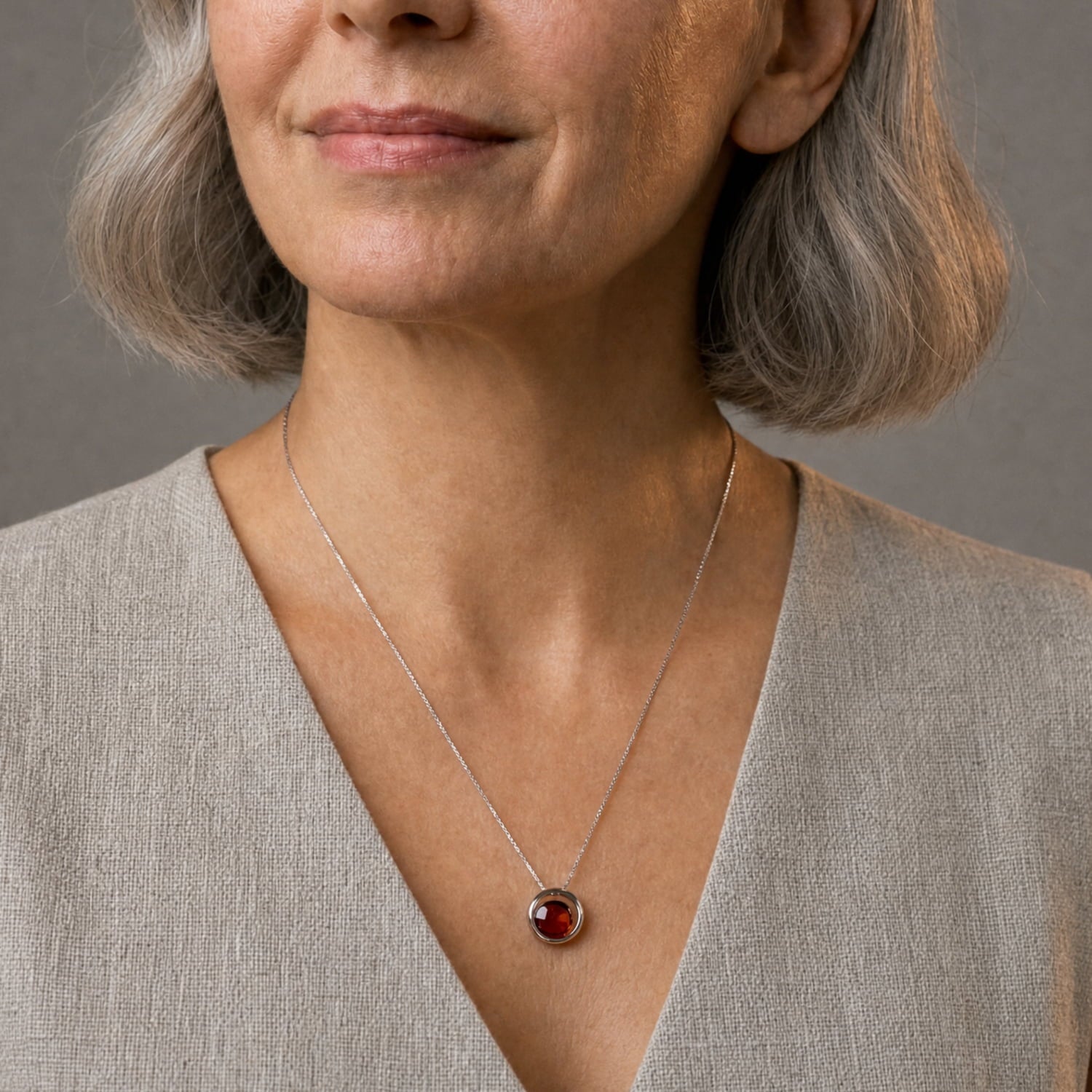 A close-up of a woman wearing a delicate silver necklace with a circular amber gemstone pendant, styled with a beige linen top against a neutral gray background.