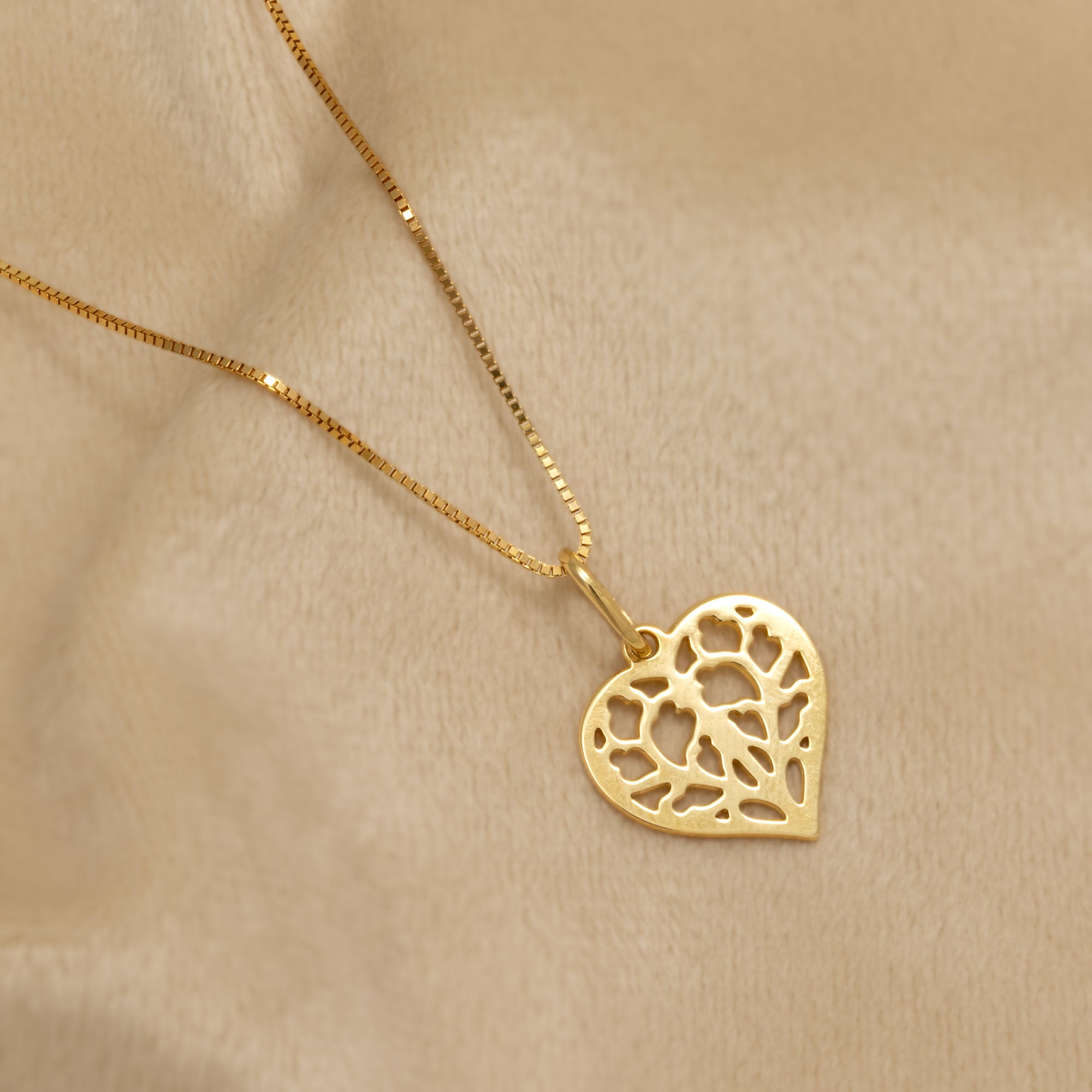 An 18ct yellow gold heart-shaped pendant featuring an intricate openwork design inspired by York Minster. It is suspended from a delicate chain for an elegant and sculptural appearance. Placed on a cream velvet cloth.