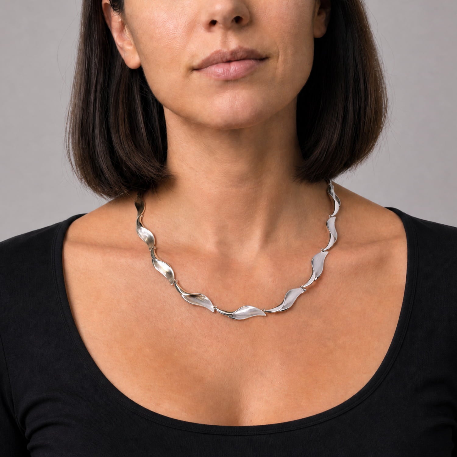 A close-up of a woman wearing a silver wavy linked necklace, paired with a black scoop-neck top against a neutral gray background.