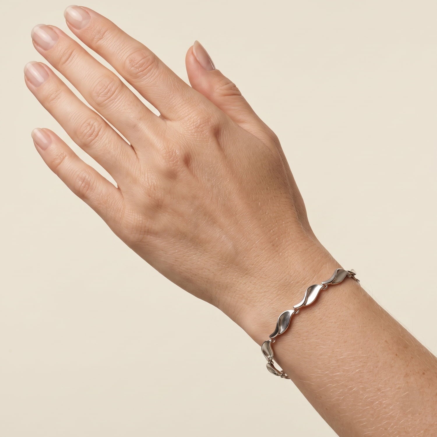 A close-up of a person's hand and wrist wearing a polished silver wavy link bracelet against a solid light beige background.
