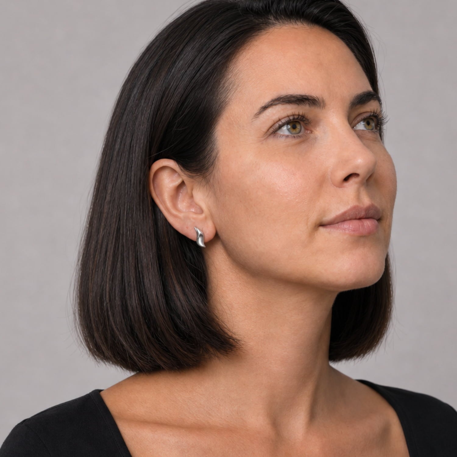 A close-up portrait of a woman with a short dark bob haircut, wearing a small silver wave-shaped stud earring, set against a plain gray background.