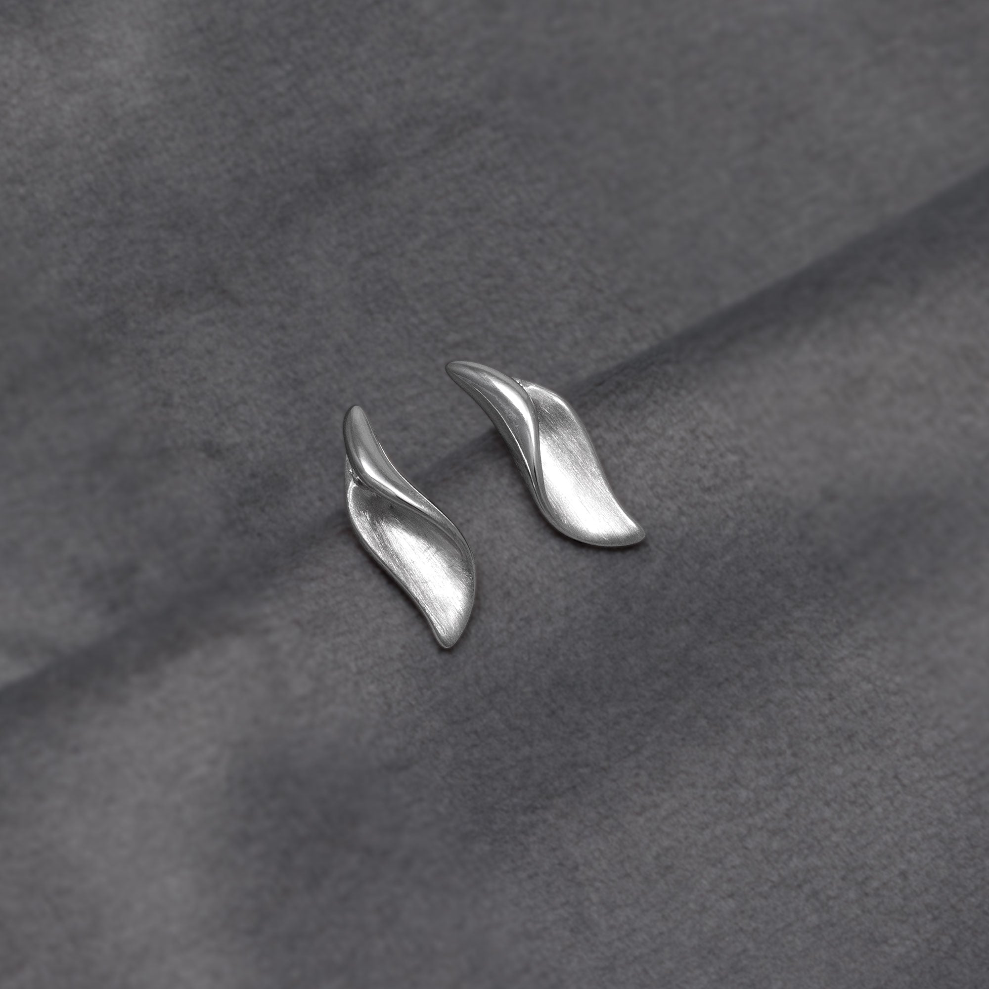 A pair of silver leaf-shaped stud earrings on a textured dark grey fabric background.