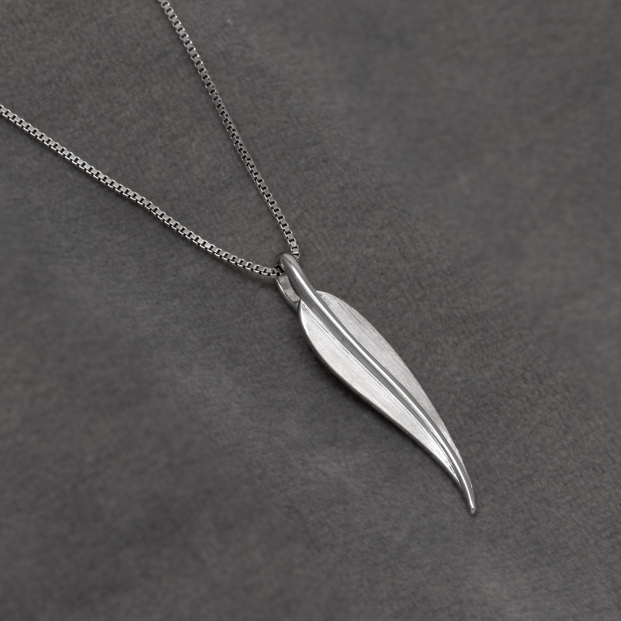 A silver pendant necklace featuring a polished, leaf-shaped charm on a delicate link chain, displayed on a textured dark grey background.