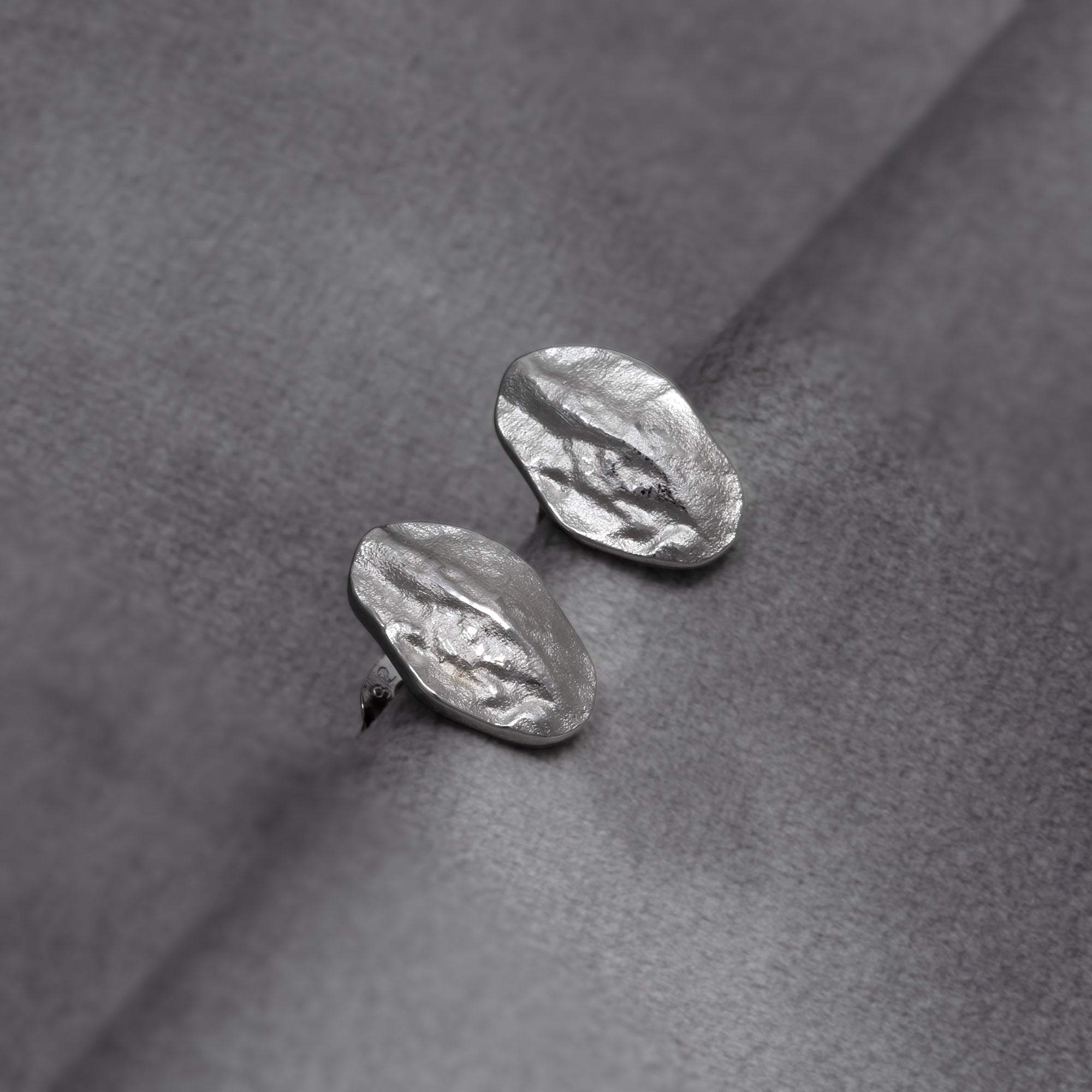 Sculptural sterling silver oval stud earrings featuring an organic, textured surface for a refined and contemporary look in British sterling silver jewellery by Azendi. Placed on a grey velvet cloth.