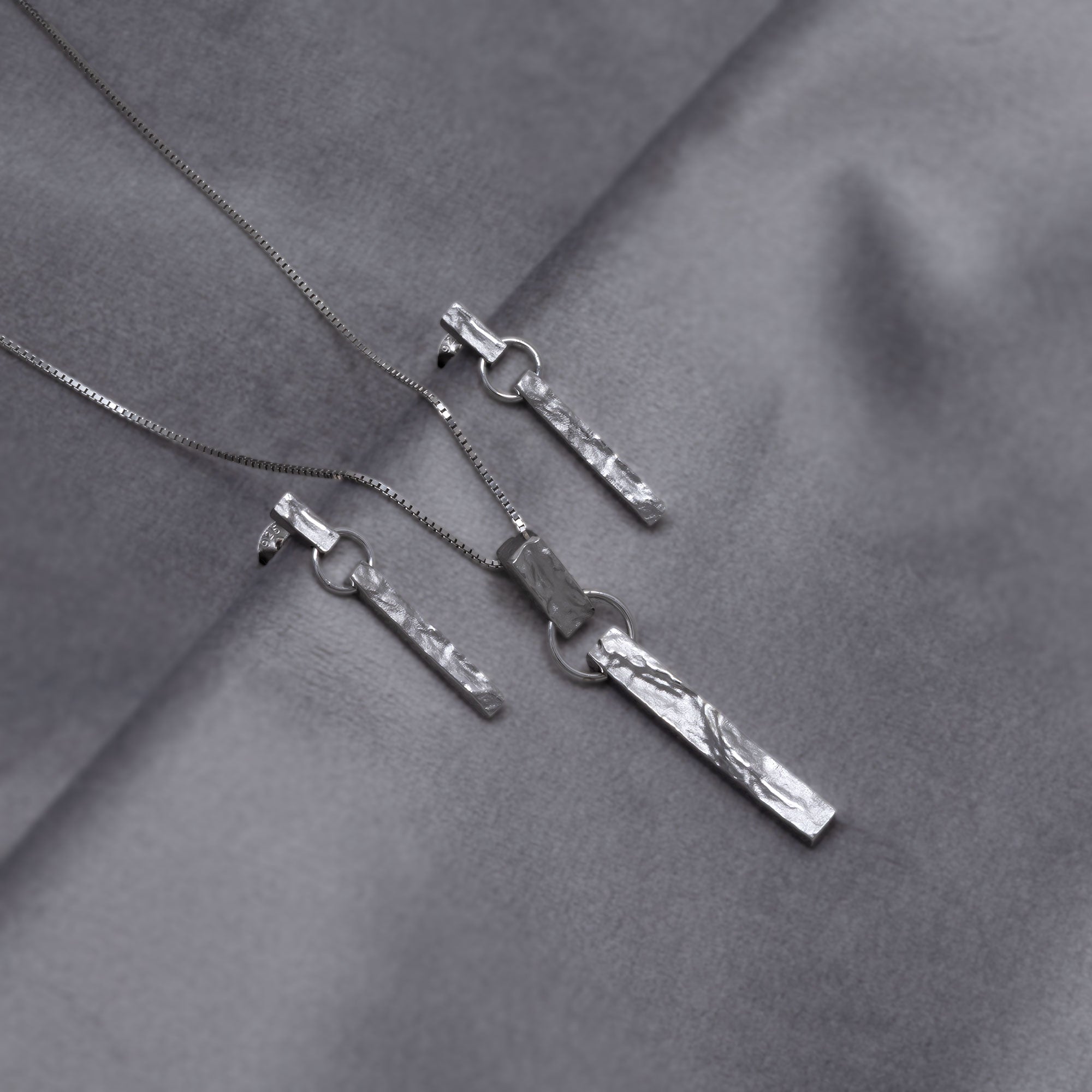 Sterling silver pendant and matching drop earrings featuring a textured bar drop design suspended from a delicate chain, showcasing a contemporary sculptural aesthetic in fine silver jewellery. Placed on a grey velvet cloth.