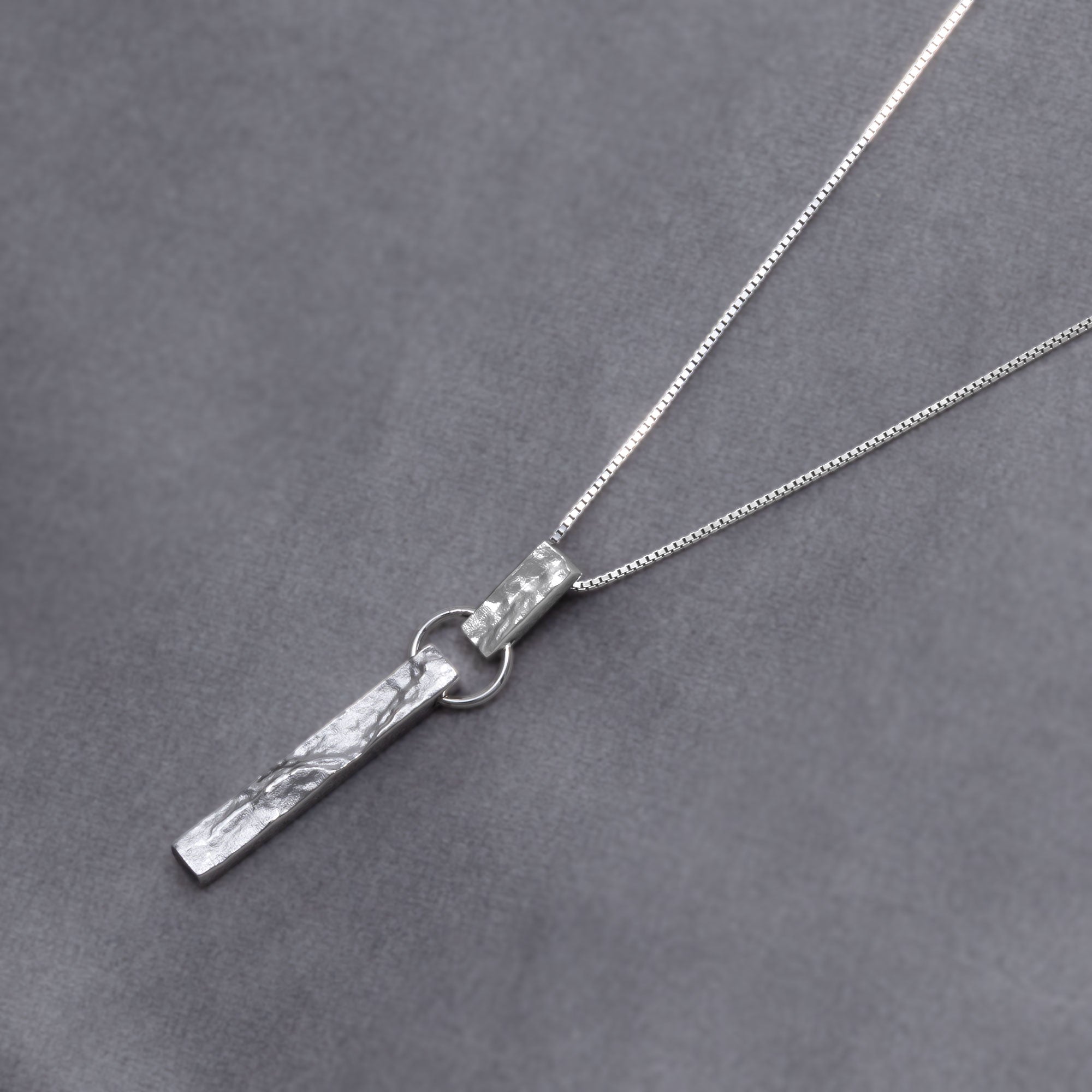 Sterling silver pendant featuring a textured bar drop design suspended from a delicate chain, showcasing a contemporary sculptural aesthetic in fine silver jewellery. Placed on a grey velvet cloth.