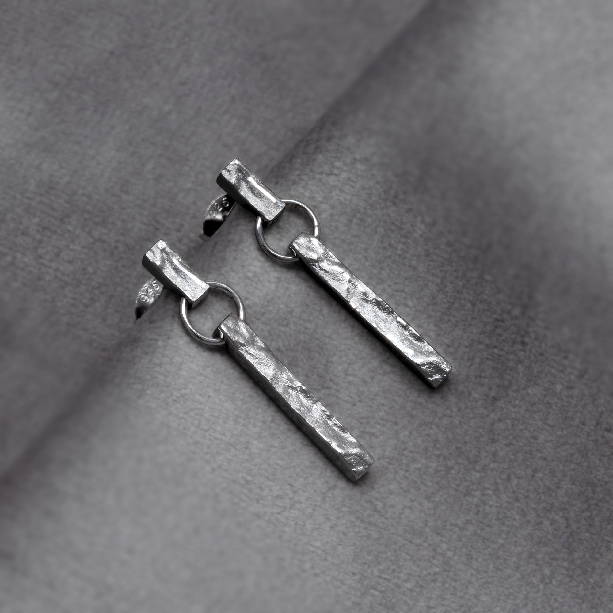 Sterling silver drop earrings featuring a textured bar drop design suspended from a delicate chain, showcasing a contemporary sculptural aesthetic in fine silver jewellery. Placed on a grey velvet cloth.