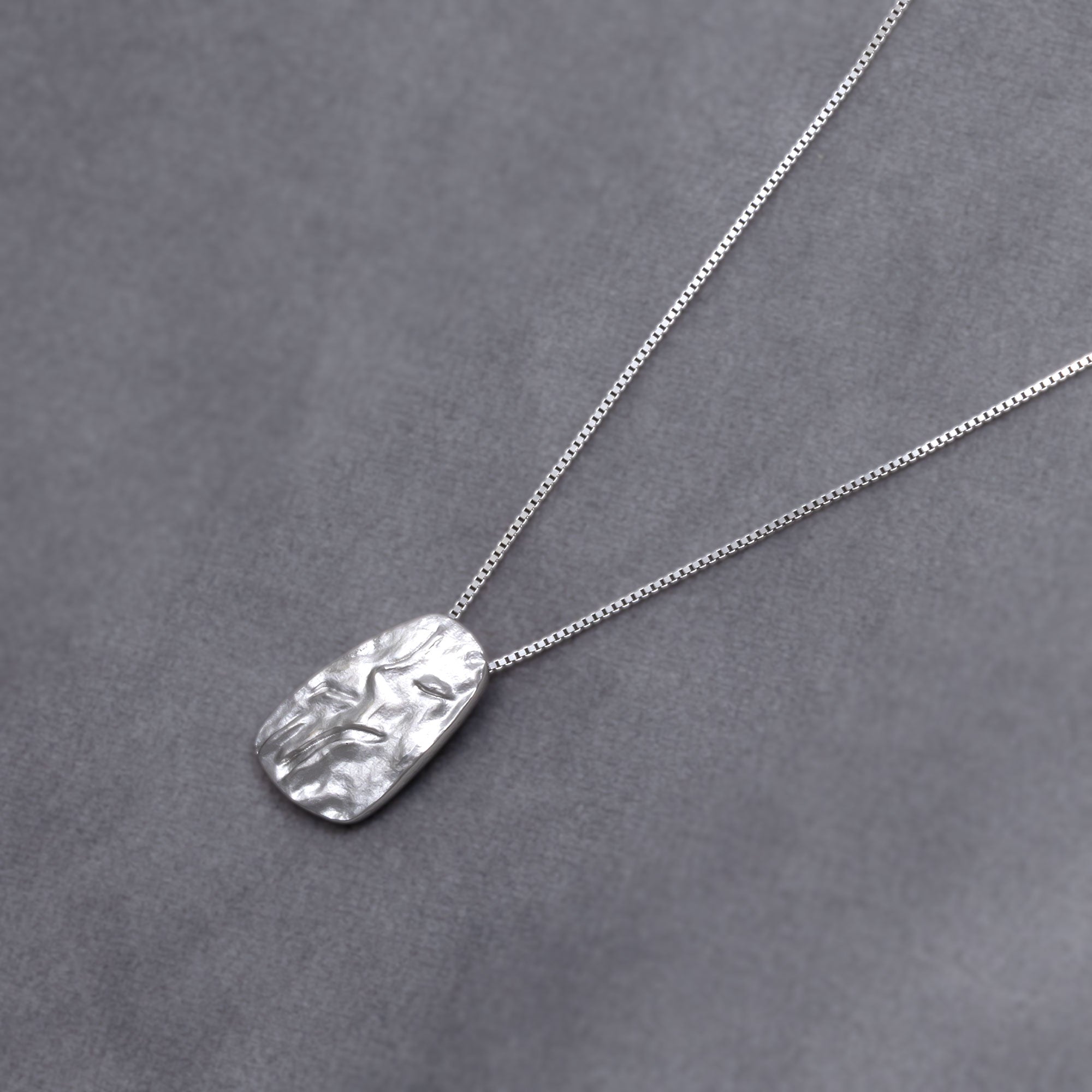Sterling silver pebble-shaped pendant with a textured, sculptural surface suspended on a delicate chain, reflecting contemporary British silver jewellery design. Placed on a grey velvet cloth.