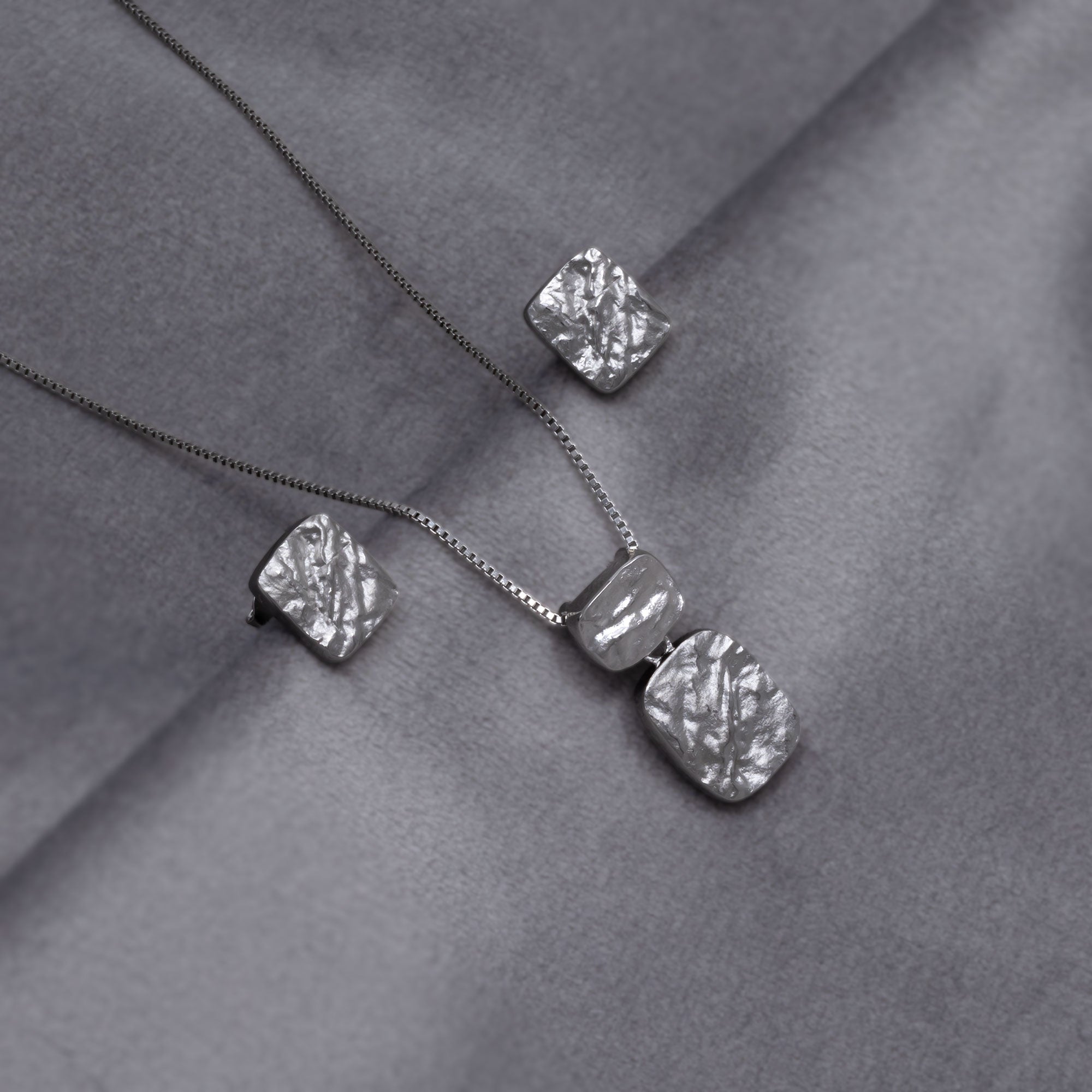 Sterling silver pendant and matching stud earrings featuring two textured rectangular elements with an organic, sculptural finish on a fine chain, embodying refined contemporary silver jewellery. Placed on a grey velvet cloth.