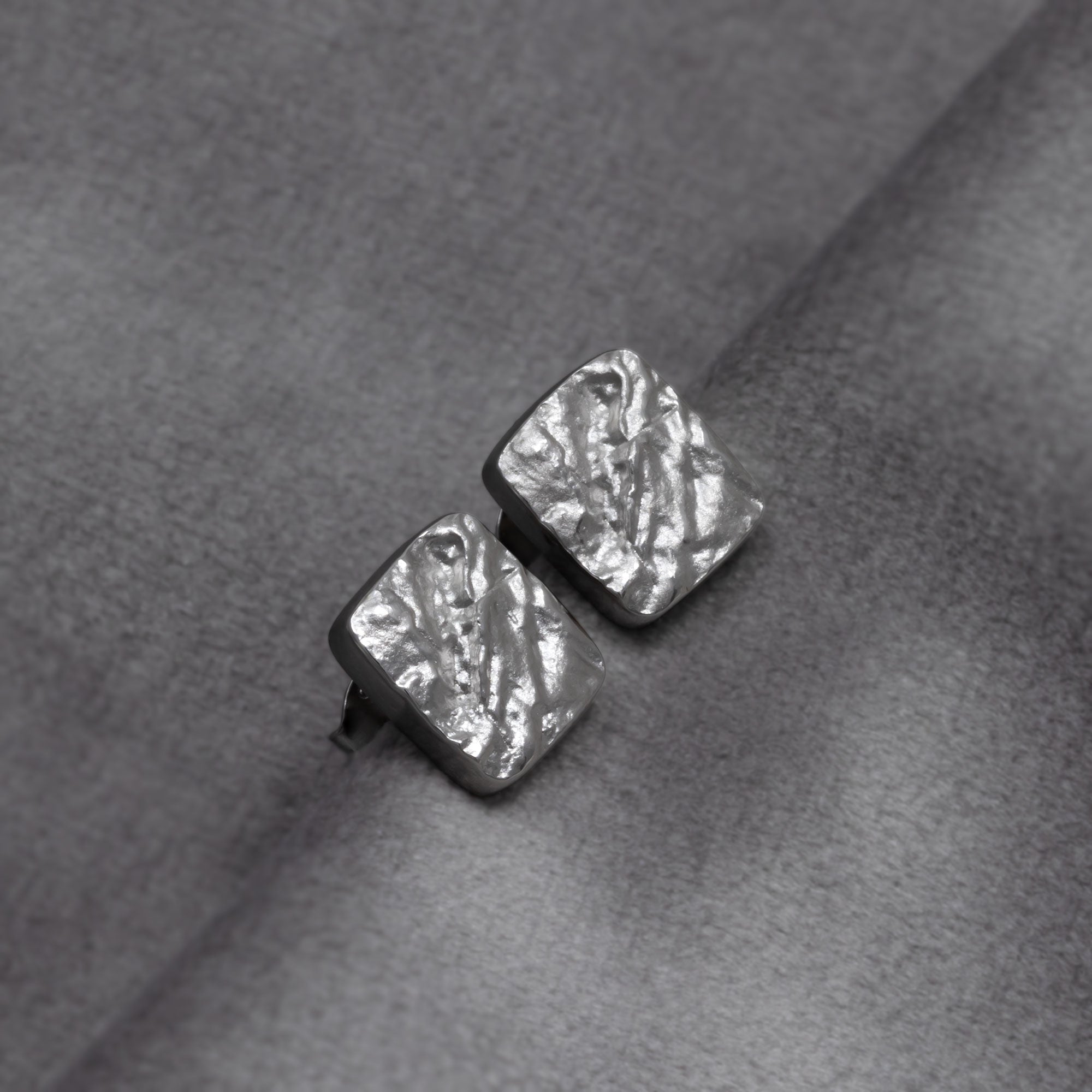 Rectangular sterling silver stud earrings with a subtle hammered texture and a polished linear detail, showcasing contemporary sculptural design in fine silver jewellery. Placed on a grey velvet cloth.