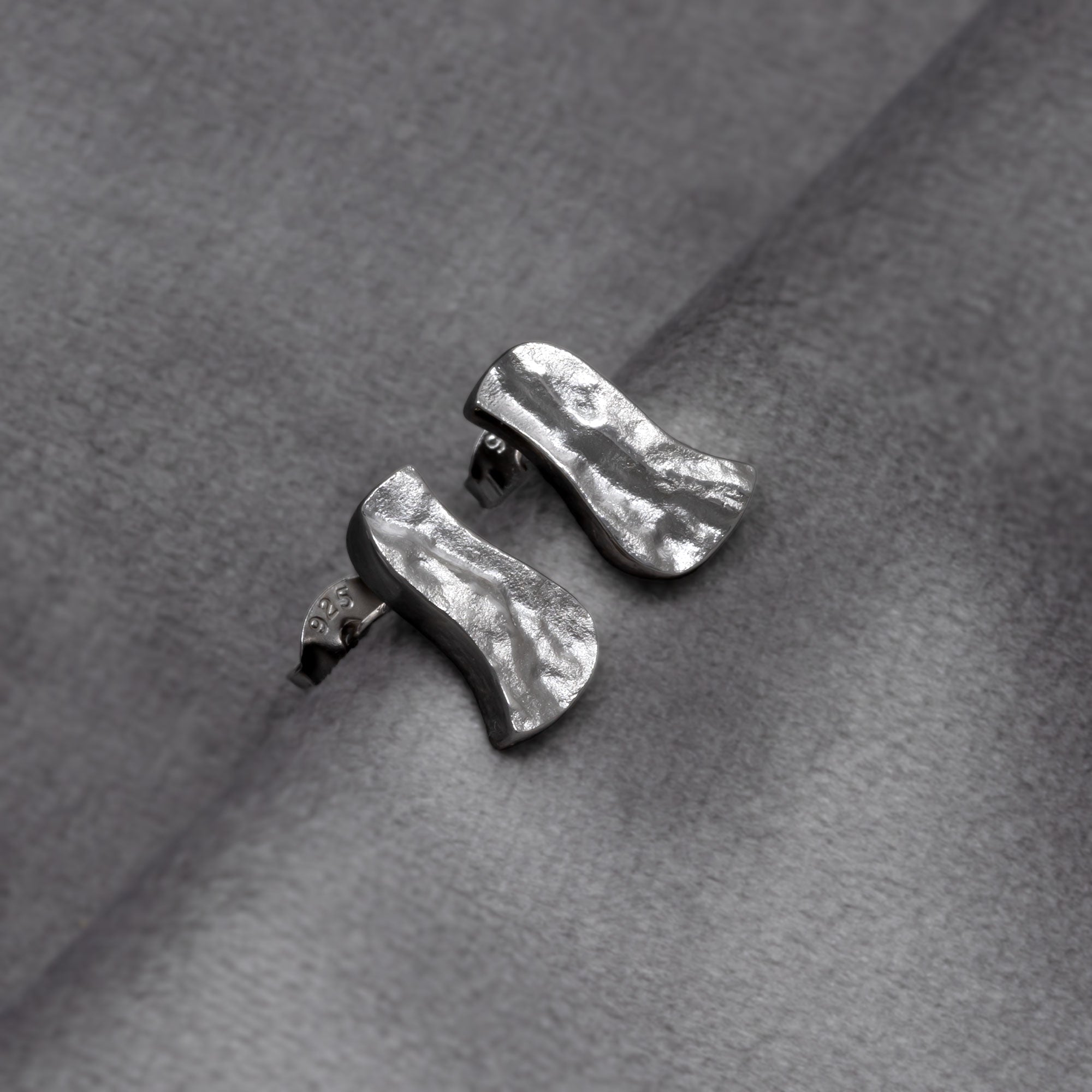 Pair of sterling silver stud earrings featuring an organic, textured wave design inspired by natural formations, embodying refined contemporary silver jewellery. Placed on a grey velvet cloth.