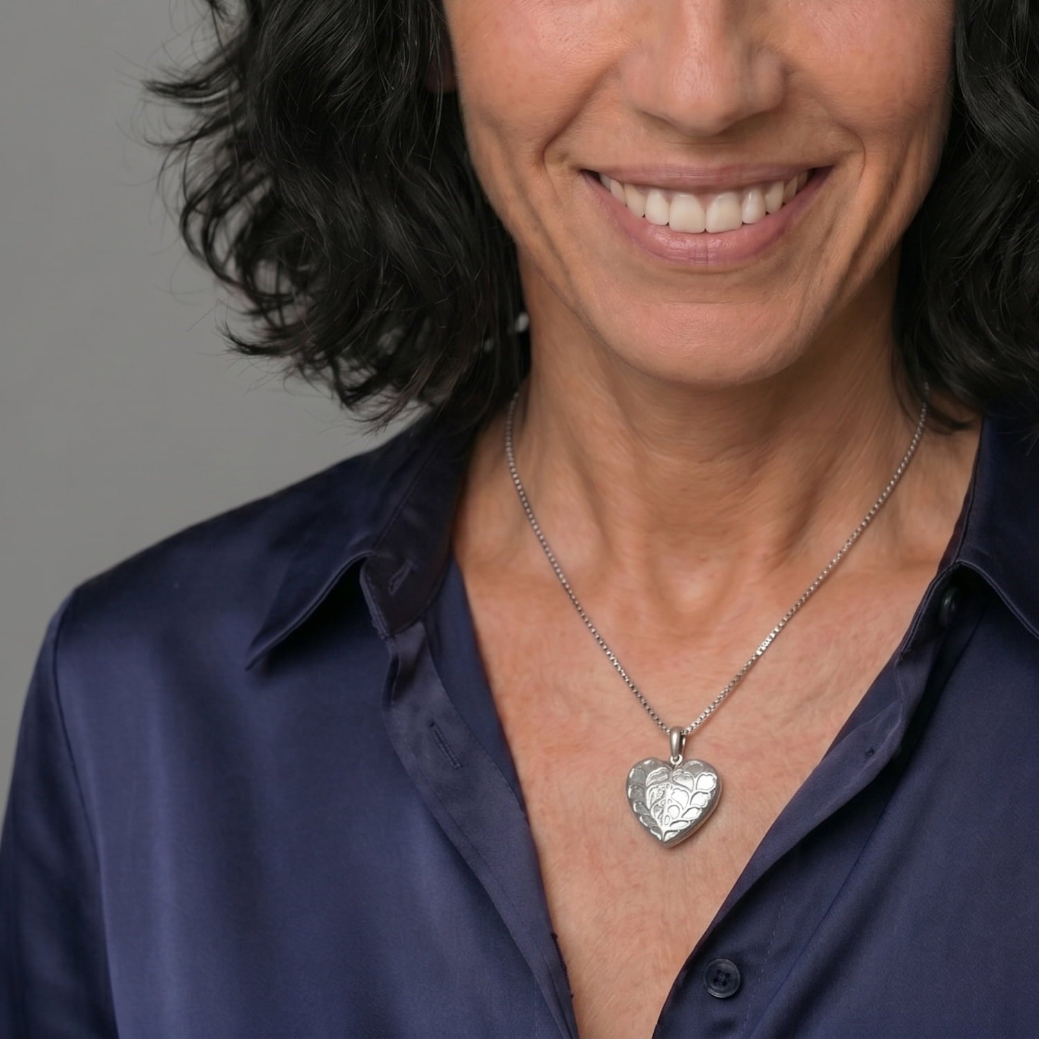 Model wears sterling silver heart-shaped locket pendant featuring intricate openwork detailing, styled with a blue silk blouse