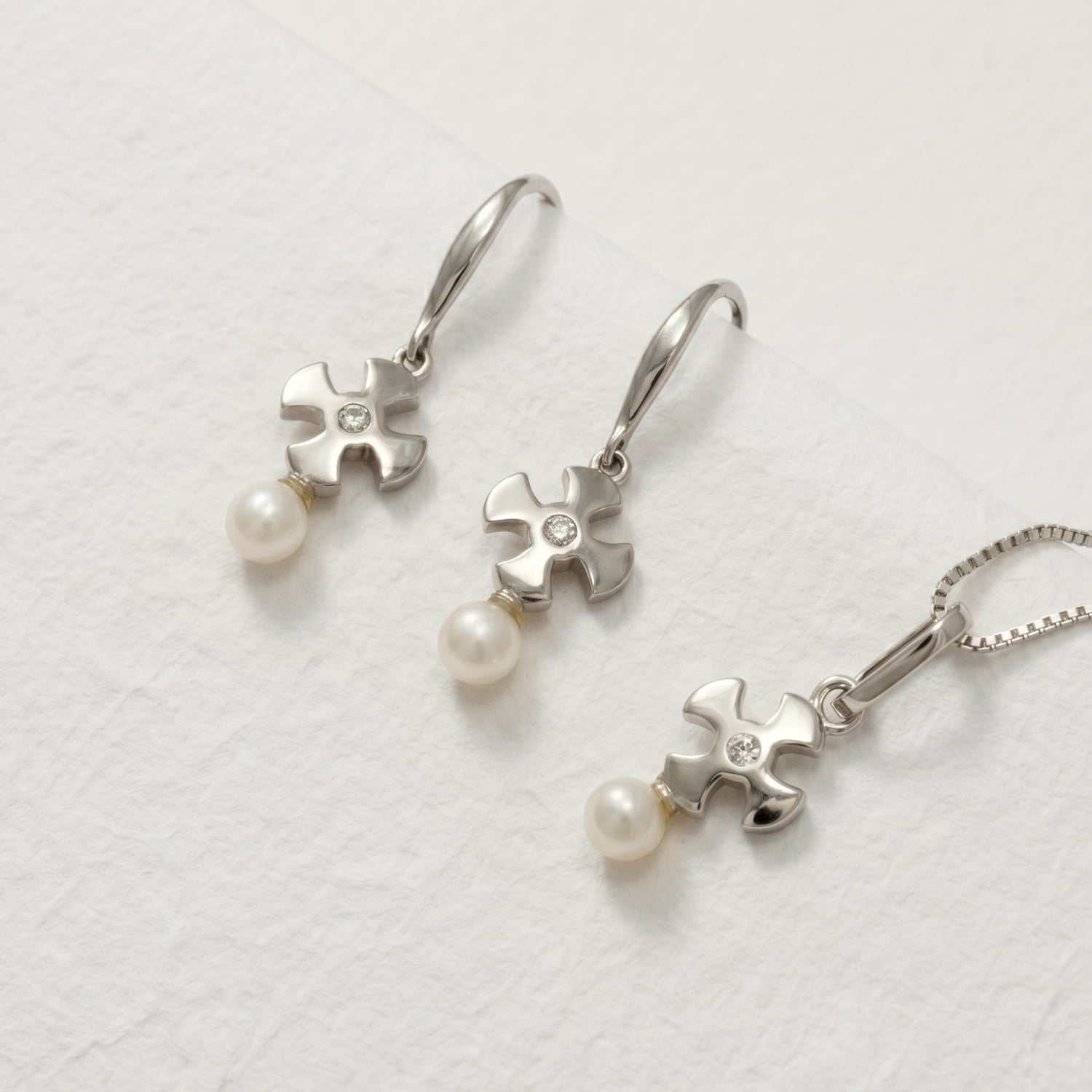 Pair of sterling silver drop earrings and a matching pendant featuring sculptural St Cuthbert crosses set with clear stones and suspended freshwater pearls on a soft beige background.Pair of sterling silver drop earrings and a matching pendant featuring sculptural St Cuthbert crosses set with clear stones and suspended freshwater pearls on a soft beige background.